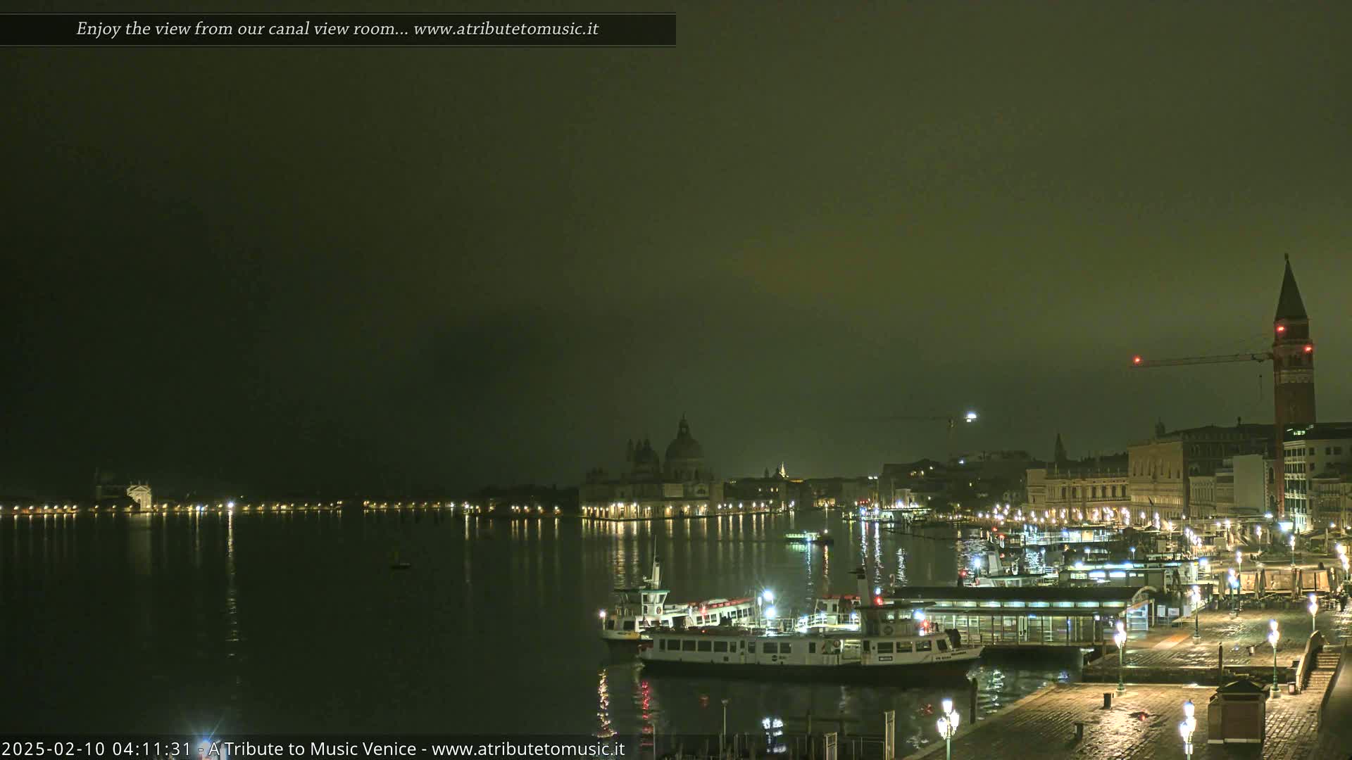 A nighttime view of a canal in Venice, with boats moored along a waterfront, under a hazy, dark sky.
