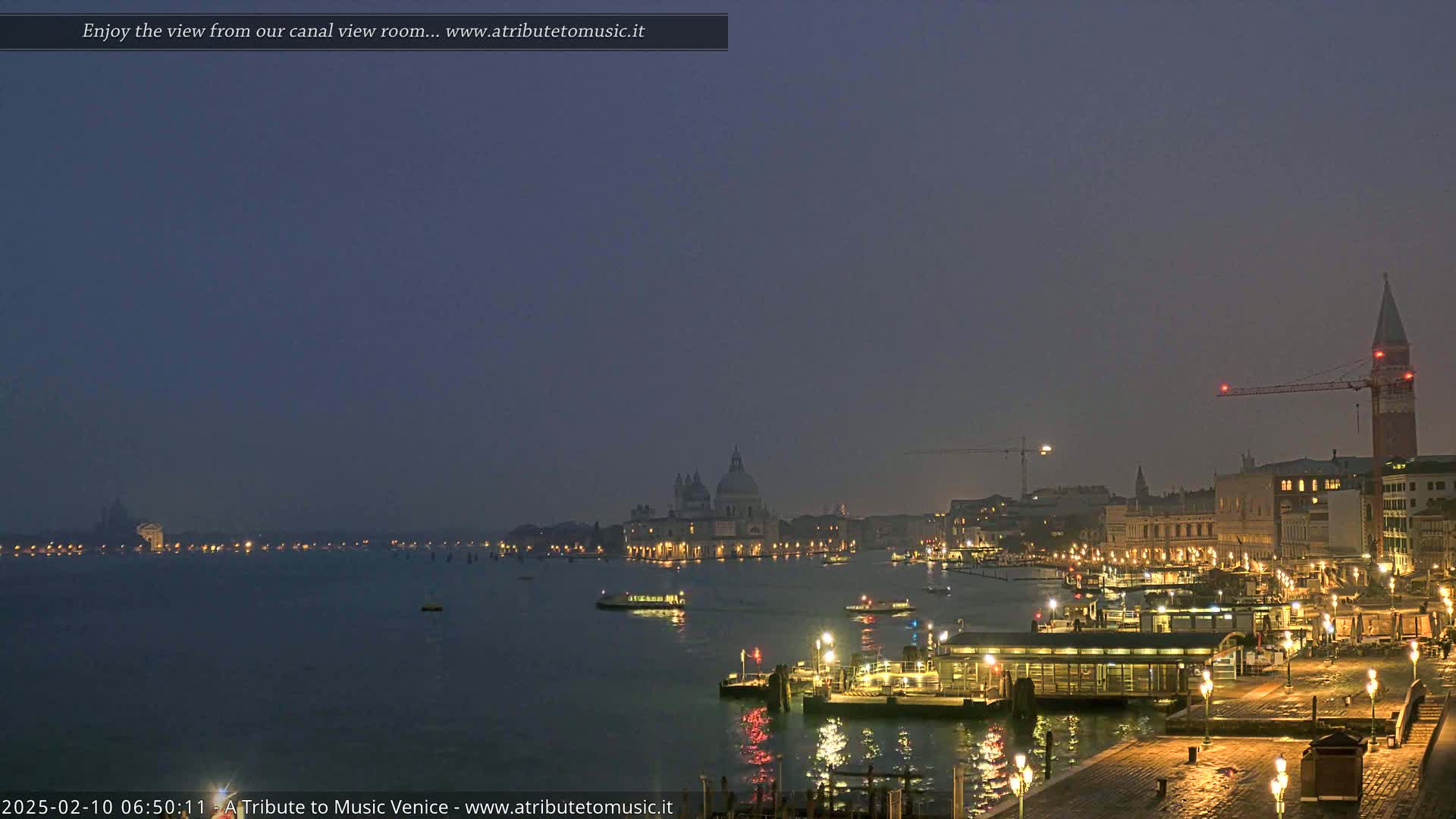 A nighttime view of Venice, Italy, shows a calm canal with illuminated buildings and boats under a hazy, dark sky.
