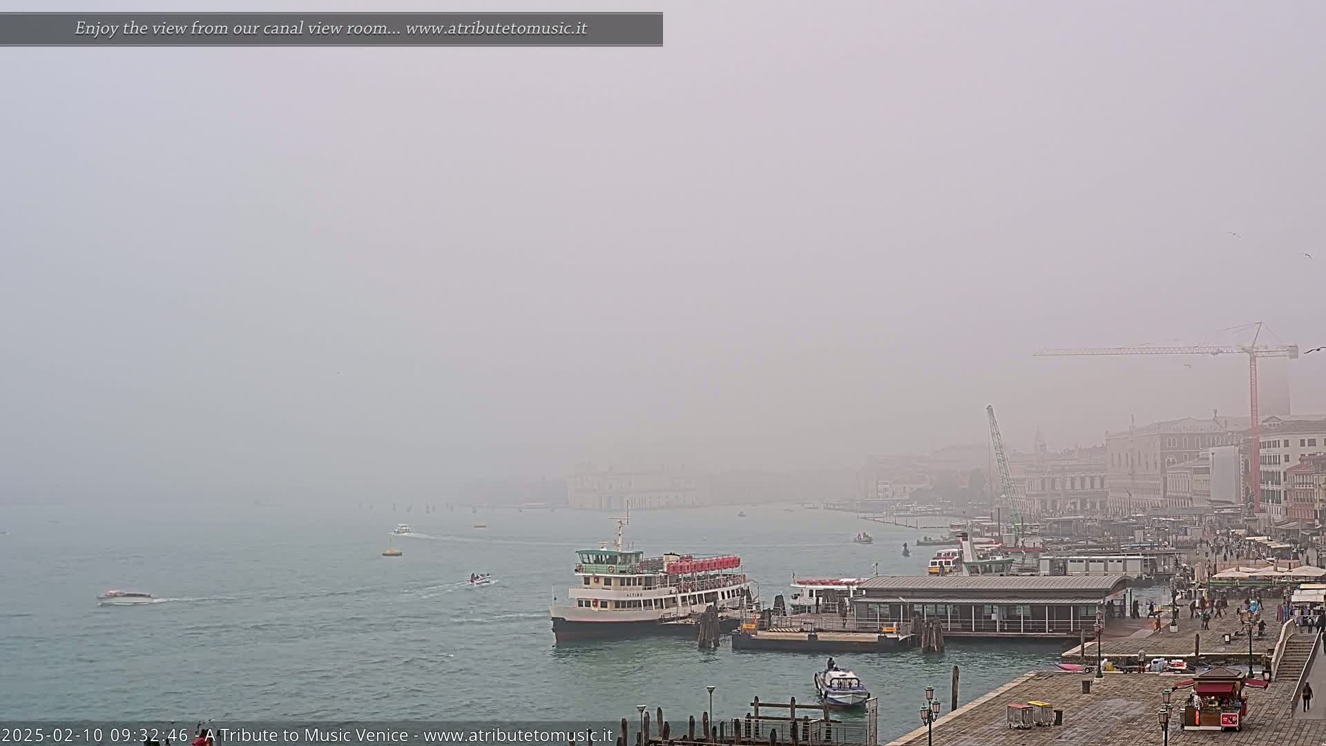 A foggy Venetian canal scene shows several boats near a dock, with buildings and a construction crane visible in the hazy distance.