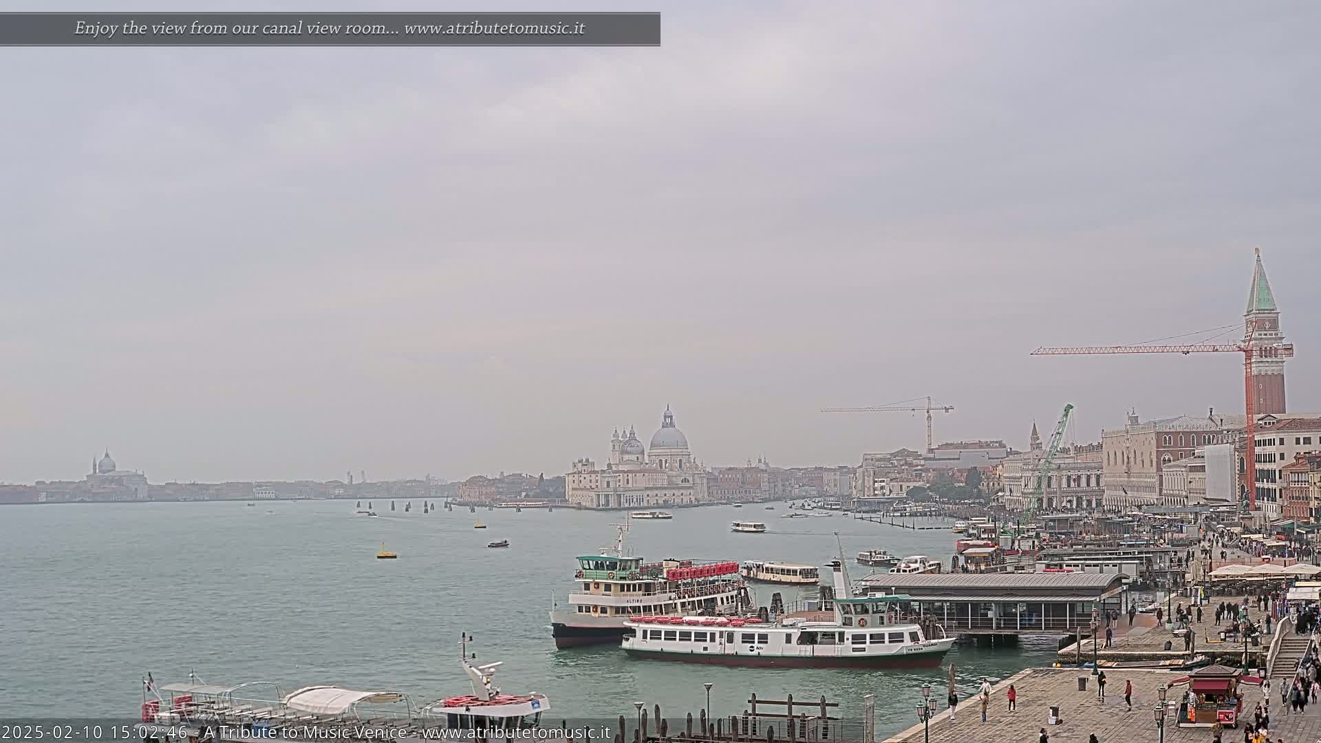 A hazy day in Venice shows a wide canal with several boats docked near a waterfront area with many pedestrians, and a distant view of the city skyline including a large church dome.