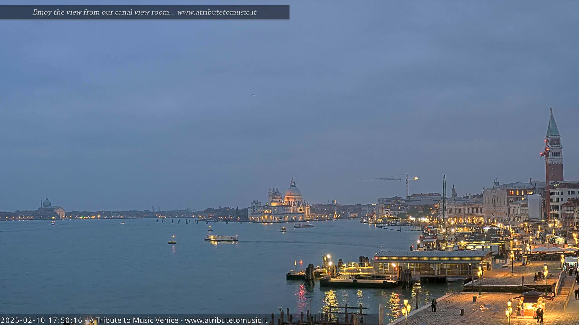 A twilight view across a calm canal shows the illuminated cityscape of Venice, including a large domed church and the bell tower of St. Mark's Square.