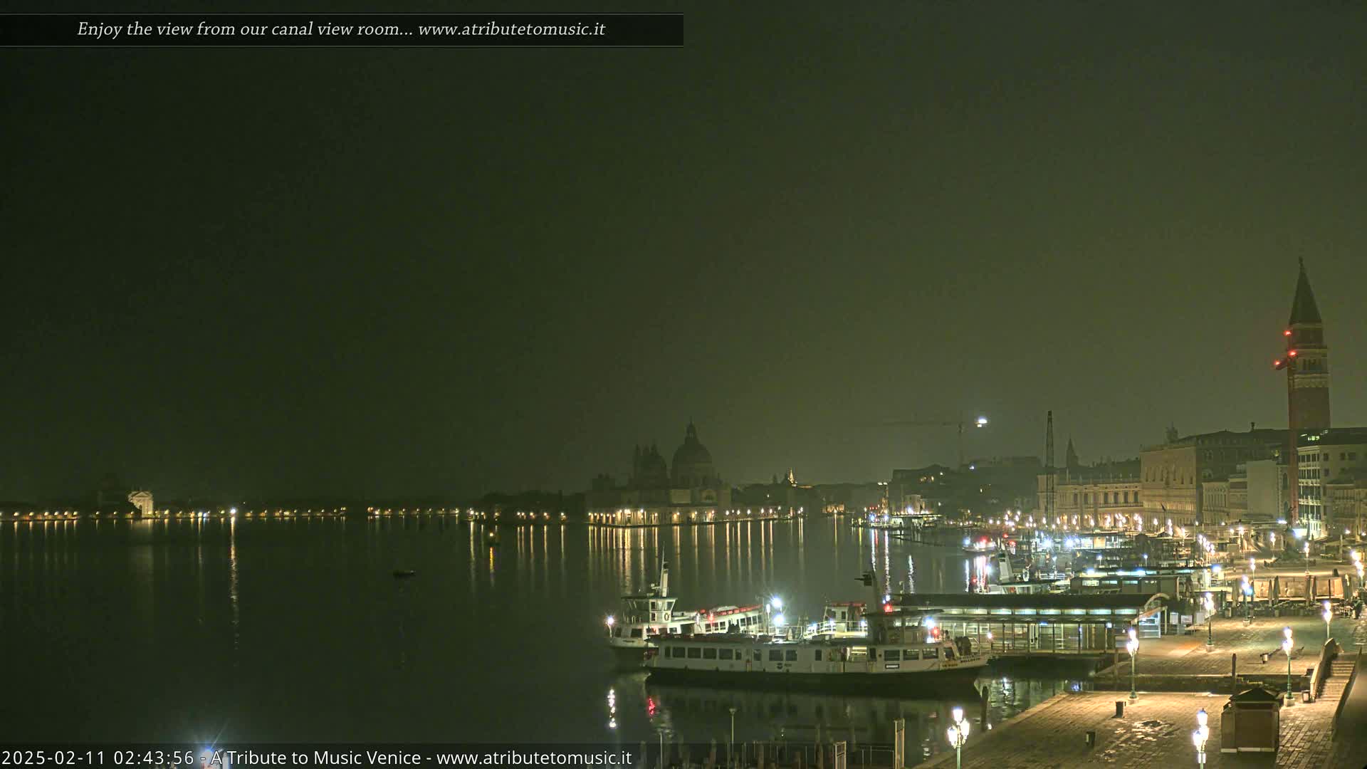 A nighttime view across a calm canal shows illuminated buildings and boats, with a hazy atmosphere.