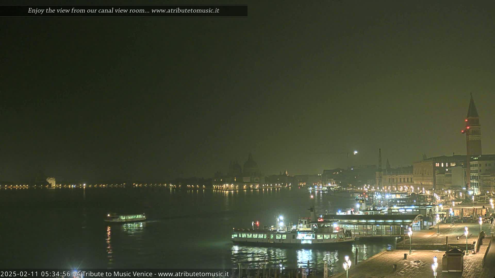 A nighttime view of a canal in Venice, shrouded in fog, shows boats docked along a waterfront with buildings and a bell tower in the distance.