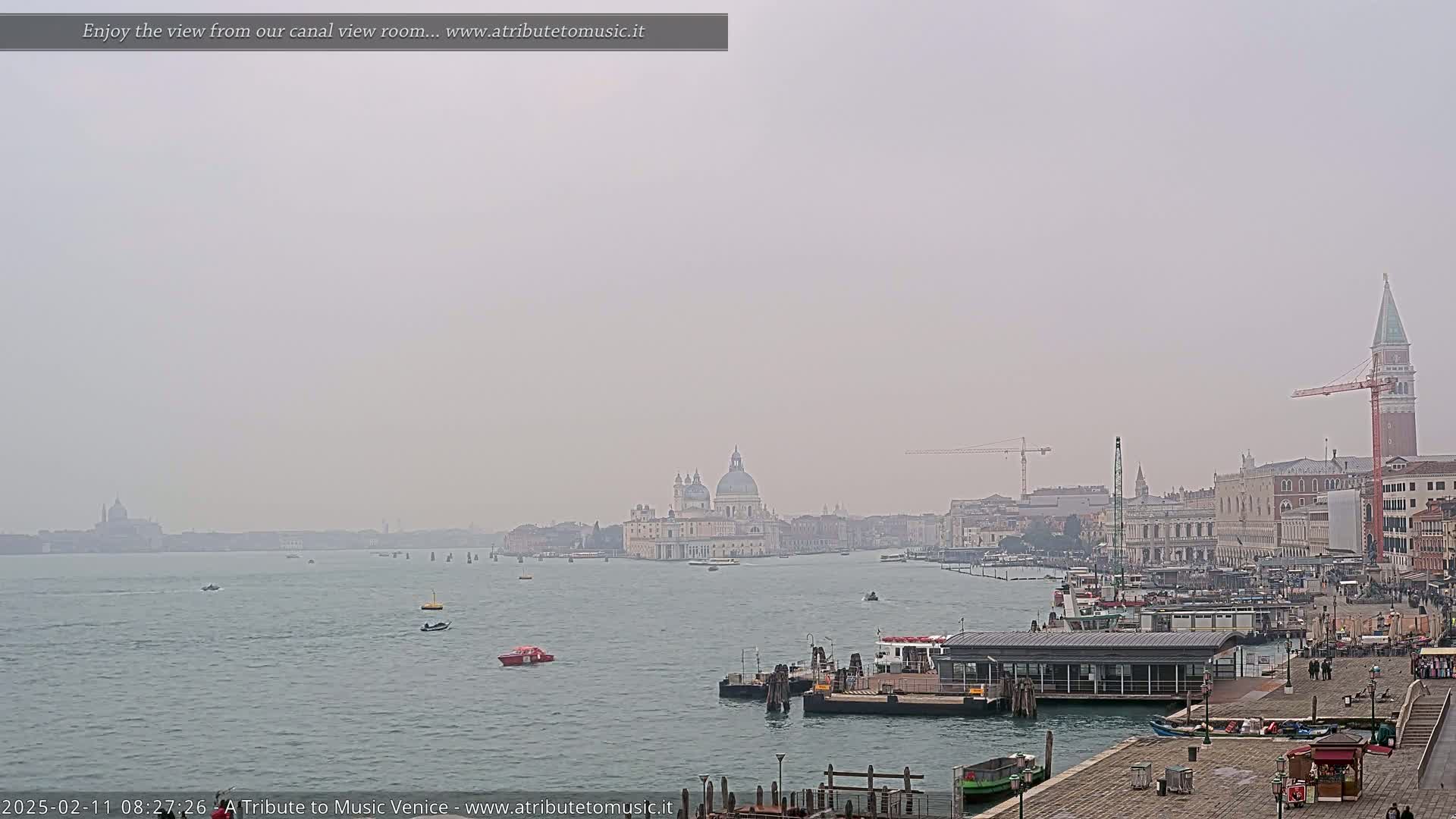 A hazy view across a canal shows a cityscape with several boats, a large building with a dome, and a tall tower in the distance.
