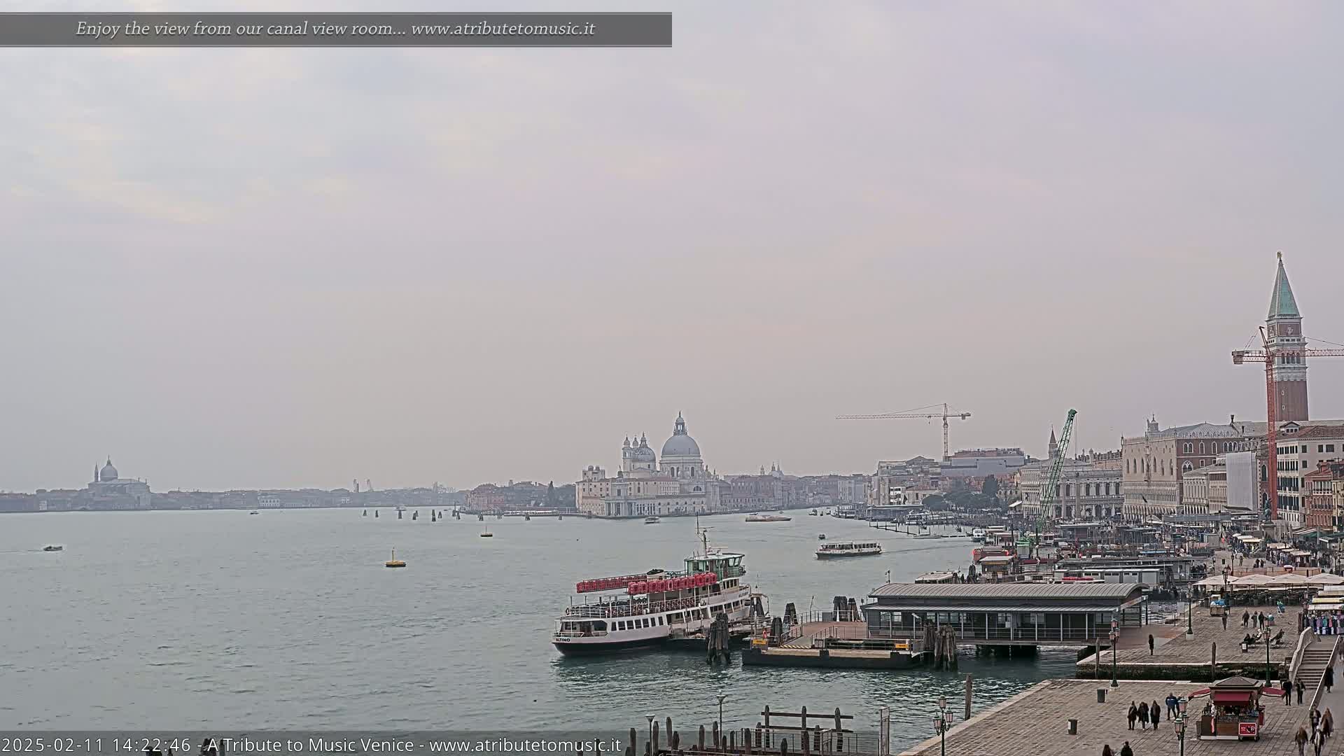A hazy, overcast day reveals a Venetian canal scene with a ferry docked near a waterfront building, a large church in the distance, and several other buildings and people along the quay.