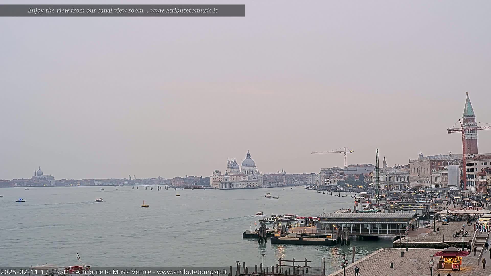 A hazy view across a calm canal shows a cityscape with a large domed church, several boats, and a tall bell tower.