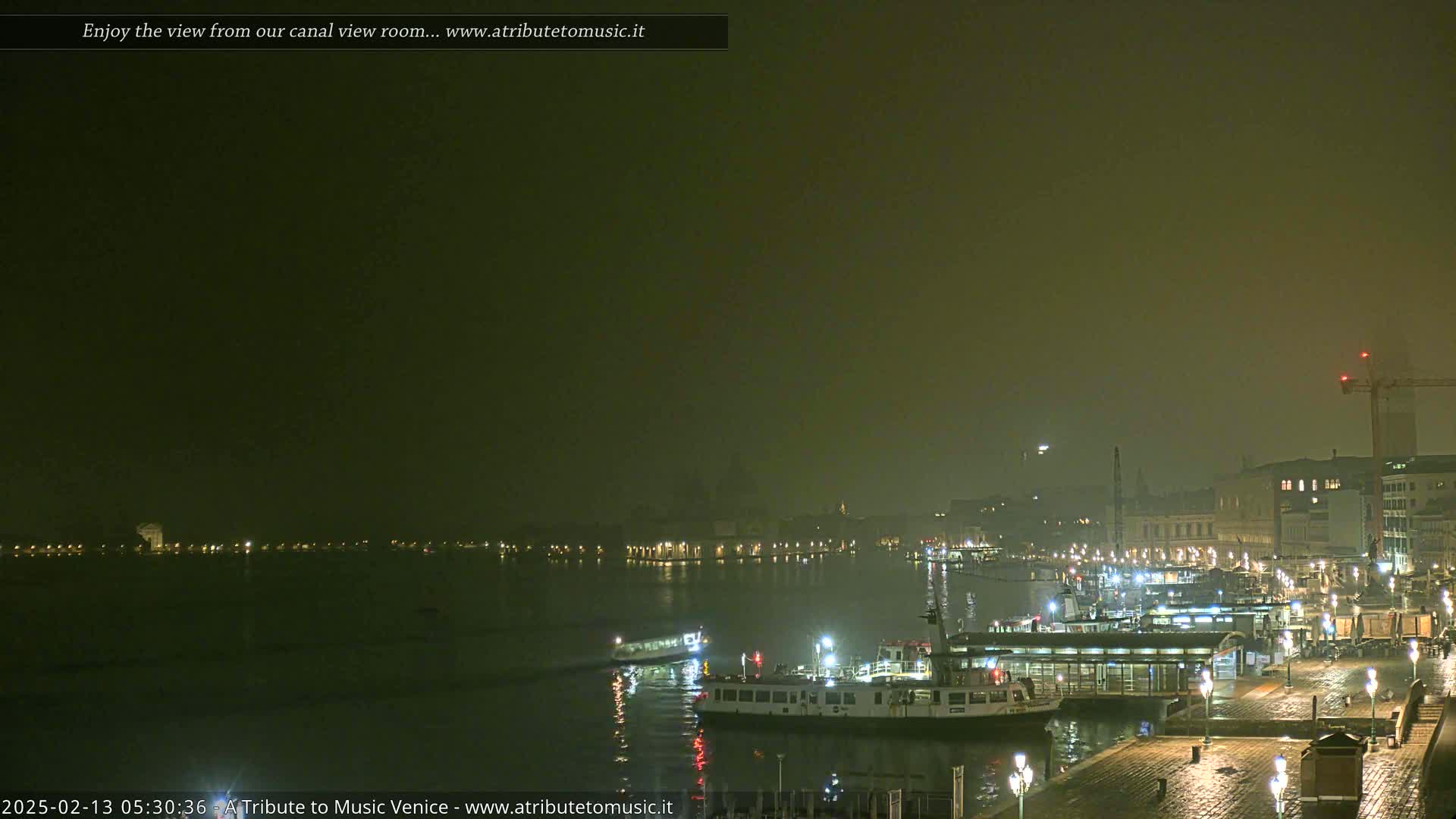 A nighttime, foggy view of a canal in Venice, showing several boats moored at a waterfront, with city lights reflecting on the water.