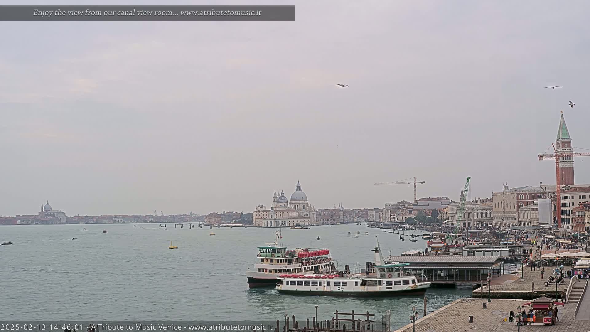 A hazy, overcast day shows a Venetian canal scene with two ferries near a dock, several boats on the water, and a cityscape including the Basilica di Santa Maria della Salute in the distance.