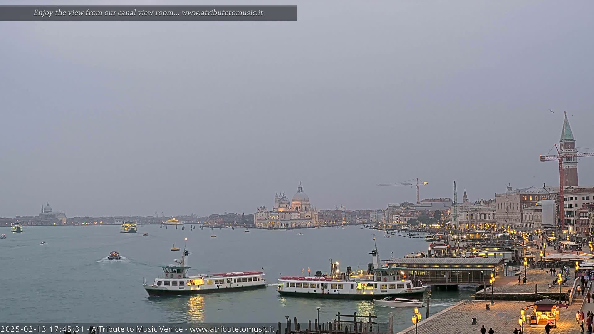 A hazy evening view over a Venetian canal shows several water buses and other boats, with the Basilica di Santa Maria della Salute and other buildings visible in the distance.