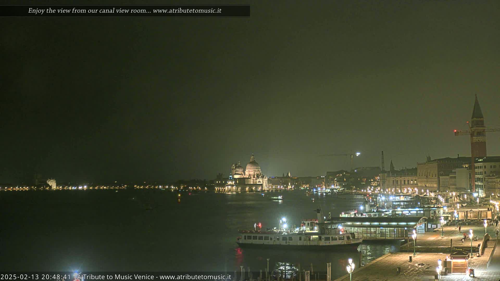 A nighttime view across a calm canal shows illuminated buildings, including a large domed structure, and several boats docked at a waterfront.