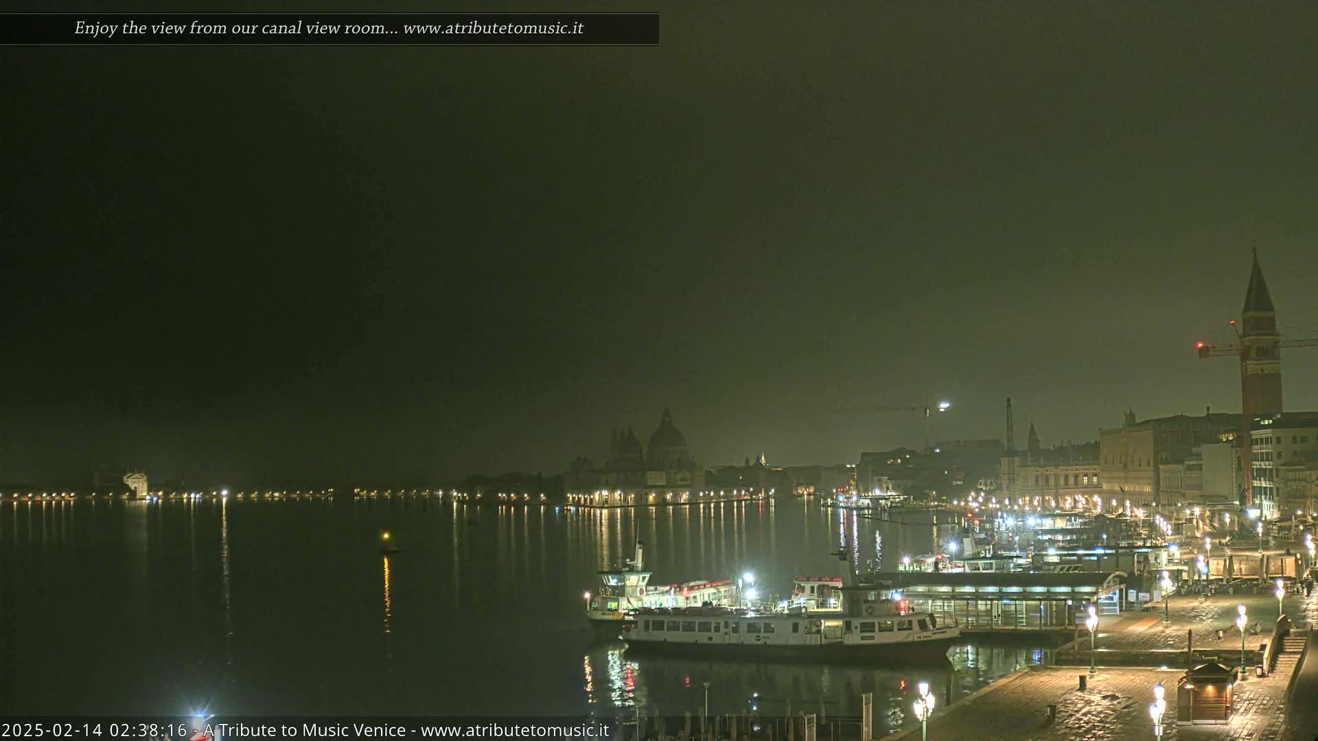 A nighttime view across a calm canal shows illuminated buildings and several boats docked at a waterfront, under a hazy, dark sky.