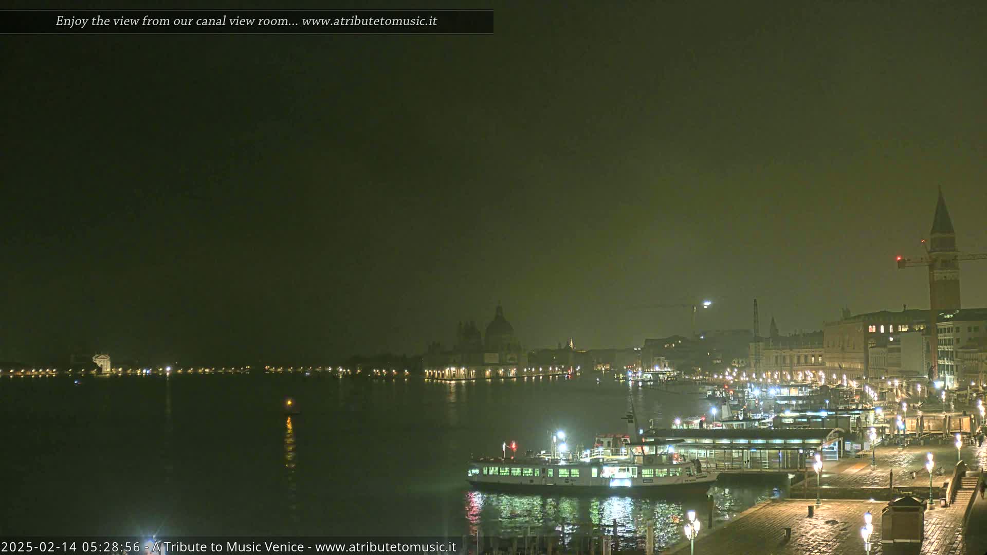 A nighttime view across a canal shows a city with illuminated buildings, boats, and a faintly visible fog or haze under a dark sky.