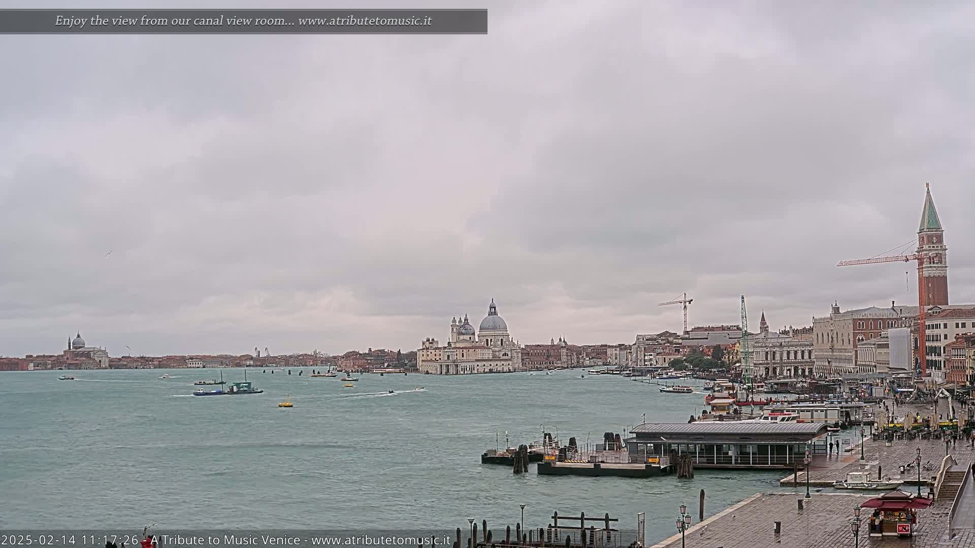 A wide, overcast view of a Venetian waterway shows several boats and buildings, including a large domed church and a tall bell tower, under a gray sky.