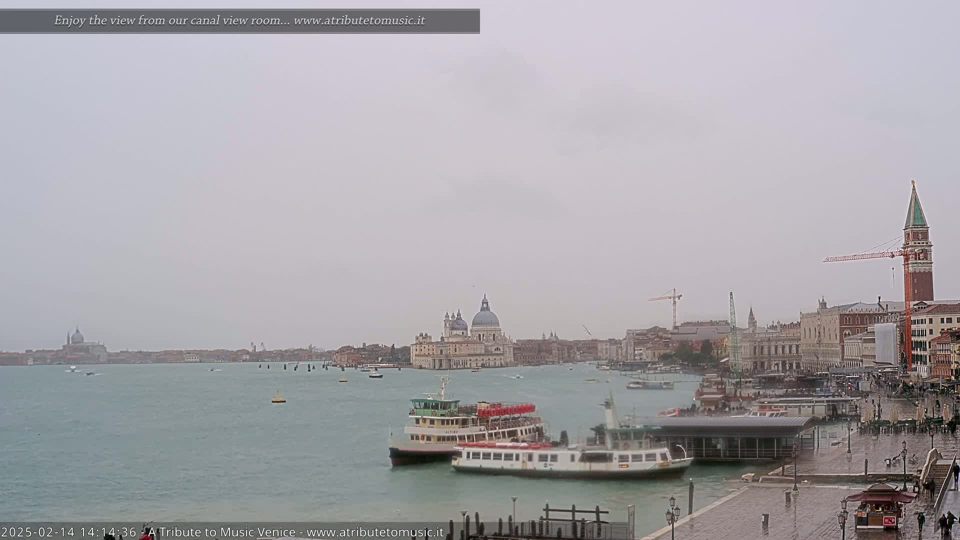 A wide shot of a waterway in Venice, Italy on an overcast day, shows several boats docked near a large building complex, with the Basilica di Santa Maria della Salute visible in the distance.