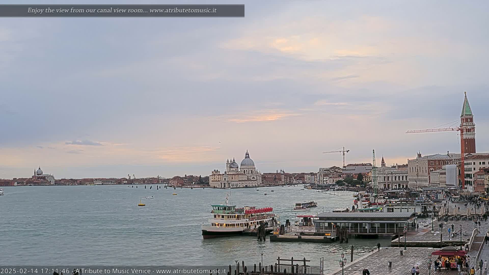 A Venetian waterway scene at sunset shows a ferry at a dock, with the Basilica di Santa Maria della Salute and other buildings visible across the water under a mostly cloudy sky.
