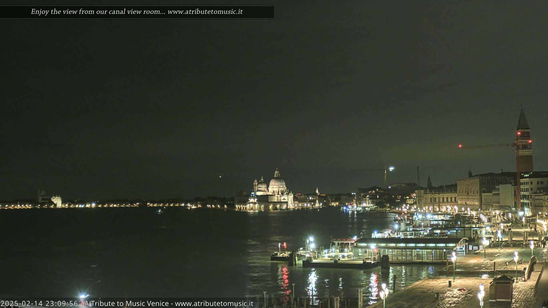 A nighttime view of a canal in Venice, showing illuminated buildings, boats, and a large church across the water under a dark sky.
