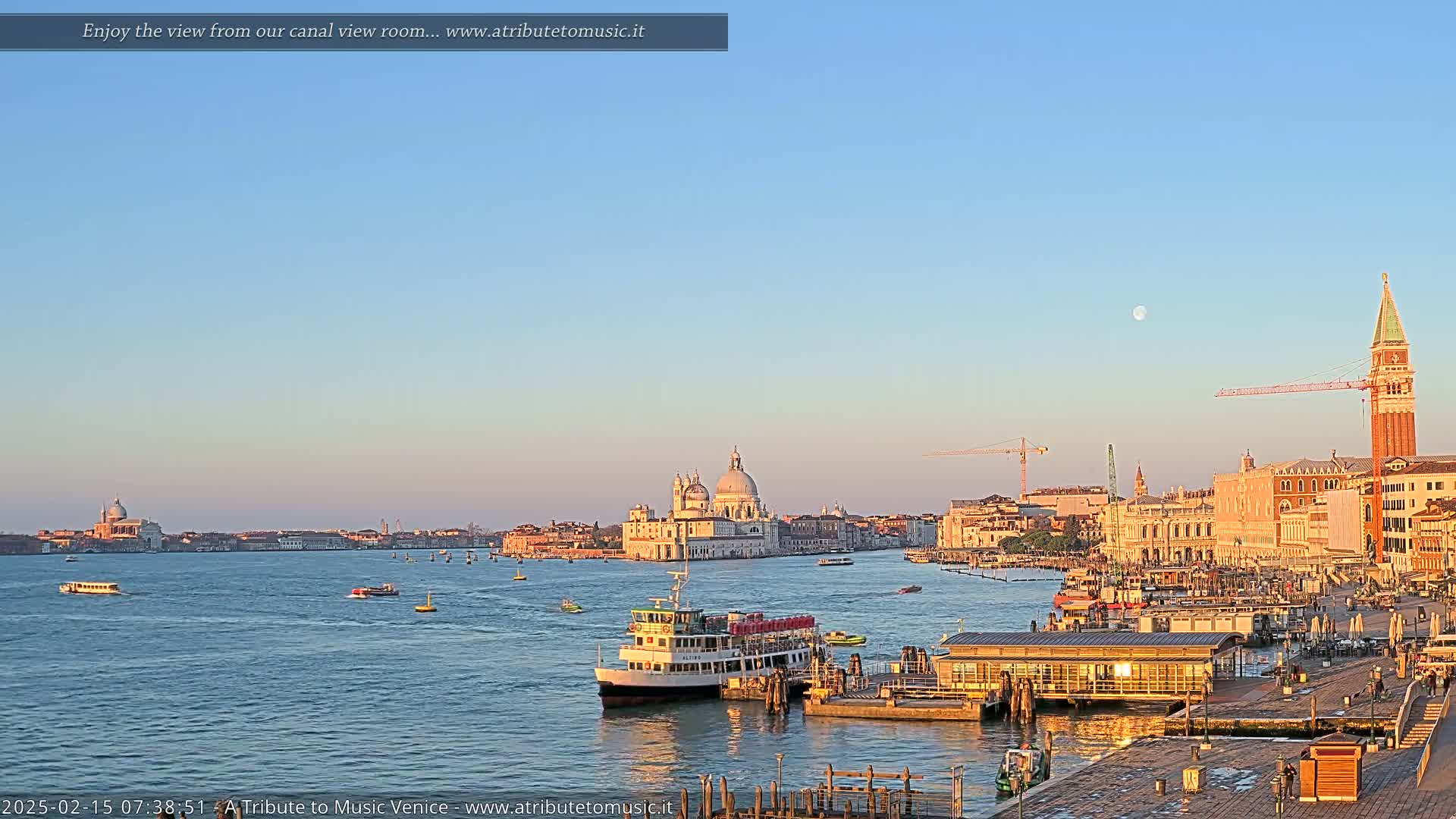 A clear, pale blue sky with a full moon hangs over a calm body of water where several boats are docked near a waterfront city, featuring a large church and a tall bell tower.