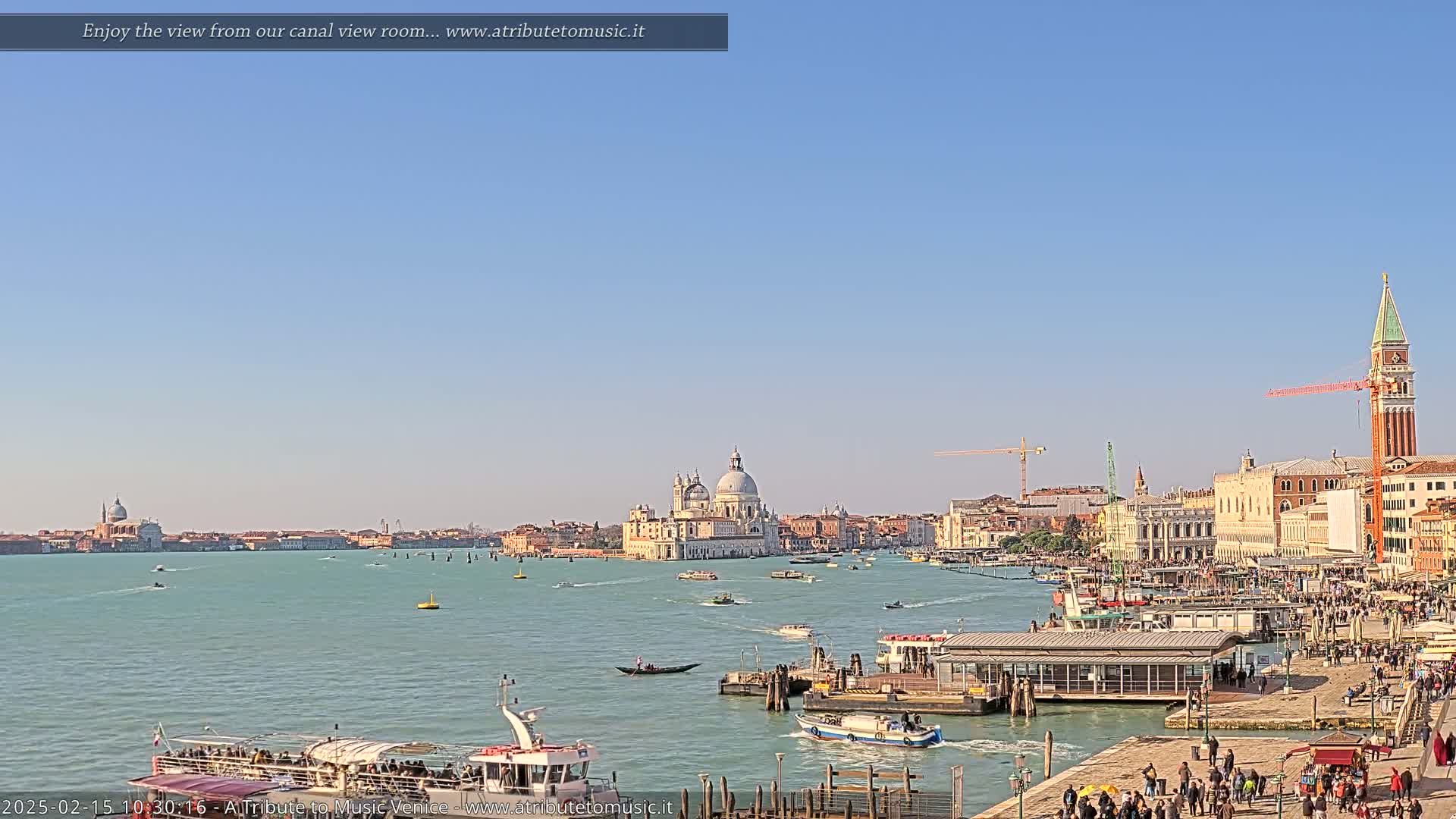 A sunny day in Venice shows a wide canal with many boats, a waterfront with people, and buildings including a large domed church and a tall bell tower in the distance.