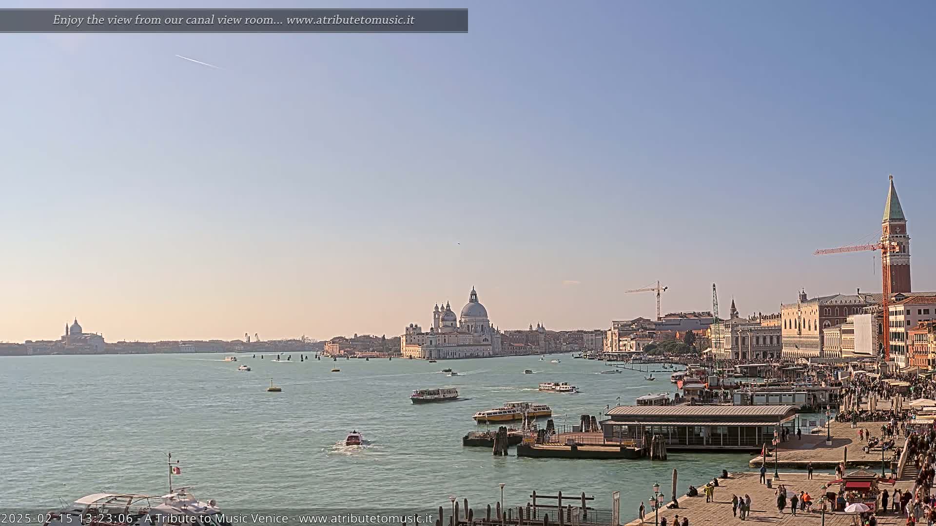 A wide, sunny view of a Venetian canal with numerous boats, a cityscape featuring a large domed church, and a bustling waterfront area.