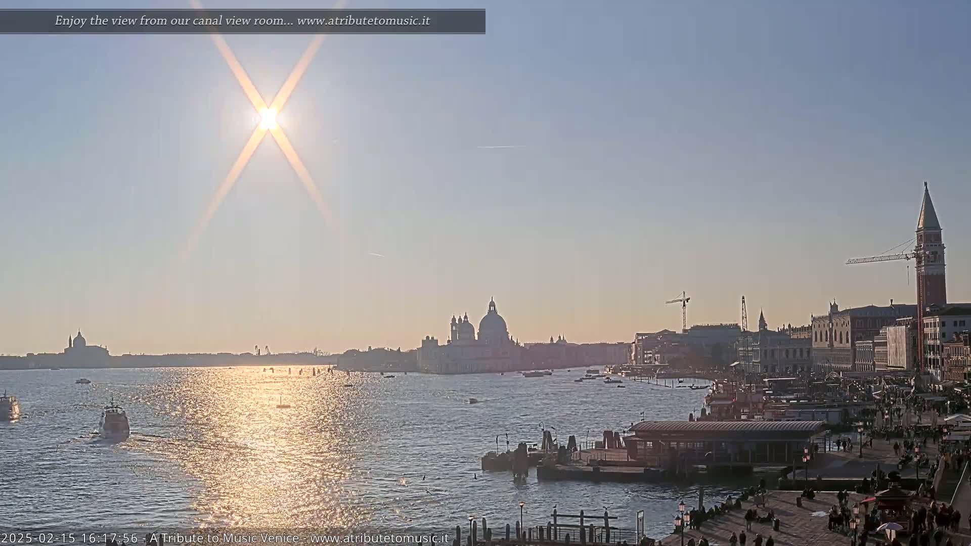 A sunny, clear day in Venice shows a canal scene with boats, buildings, and people along the waterfront, with the sun shining brightly in the sky.