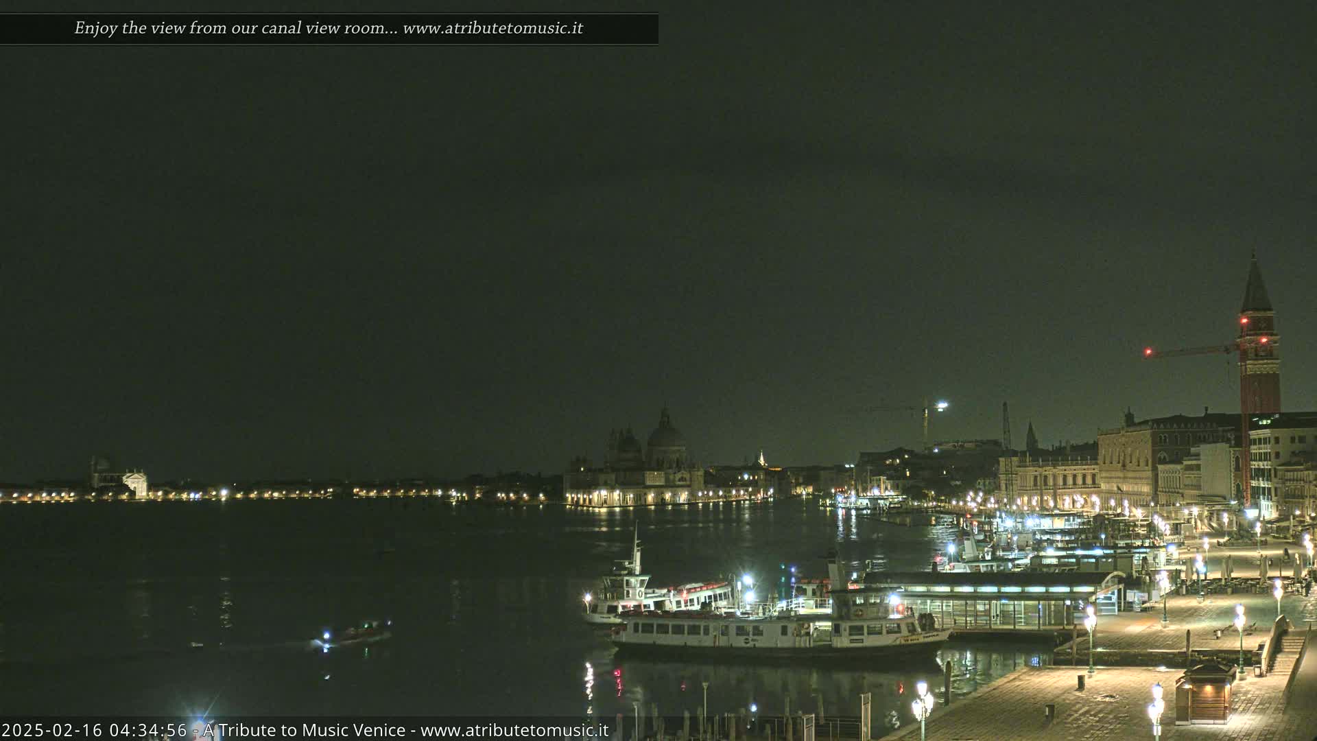A nighttime view of a calm canal in Venice, showing illuminated buildings, boats, and a sparsely populated waterfront under a dark, cloudy sky.
