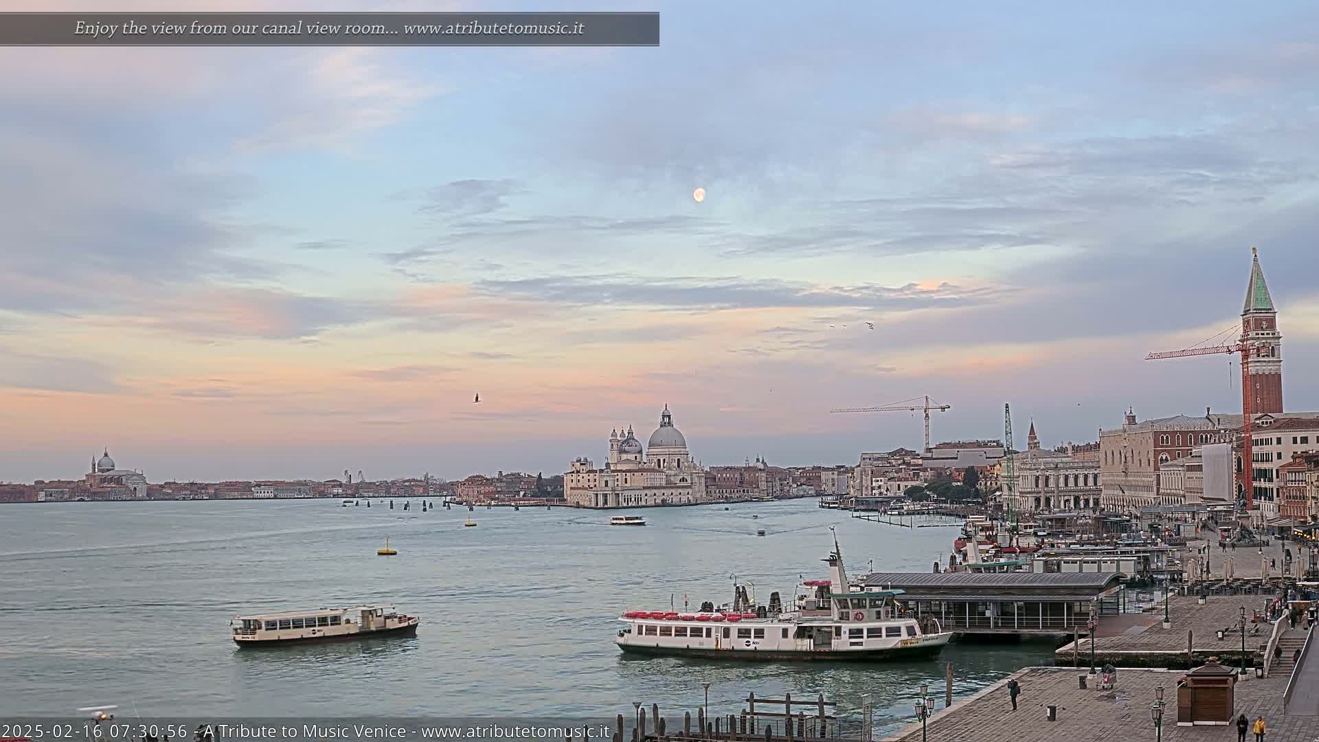 A tranquil Venetian cityscape at dawn, with a pale sky, a full moon, and several boats on calm water.
