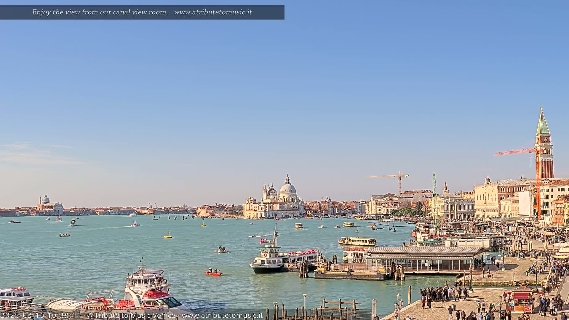 A sunny day in Venice shows a wide canal with numerous boats, a waterfront bustling with people, and a cityscape featuring the Basilica di Santa Maria della Salute and St. Mark's Campanile.