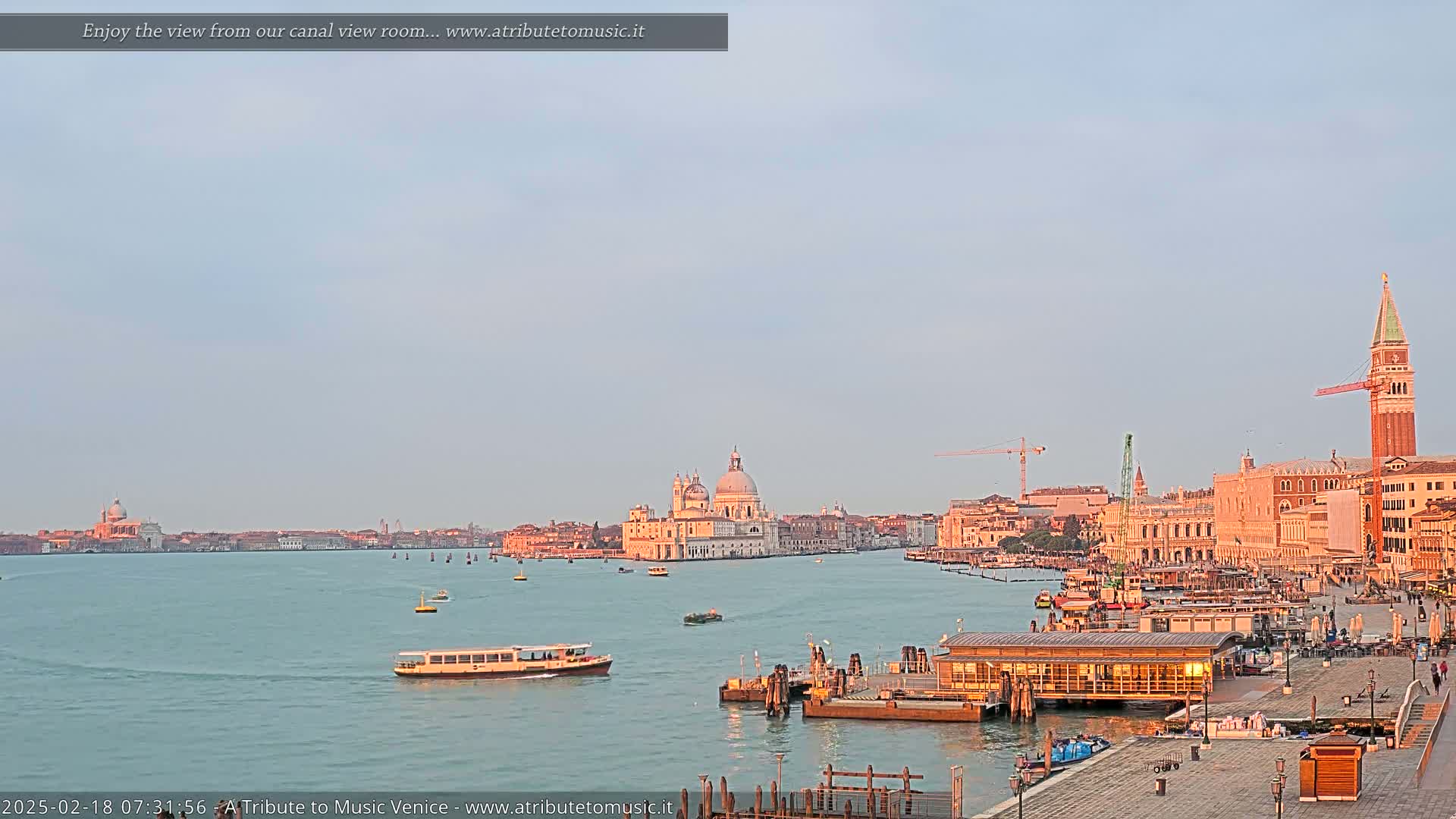 A tranquil Venetian canal scene at sunrise shows a calm waterway with boats and buildings, including the Basilica di Santa Maria della Salute, in the distance.