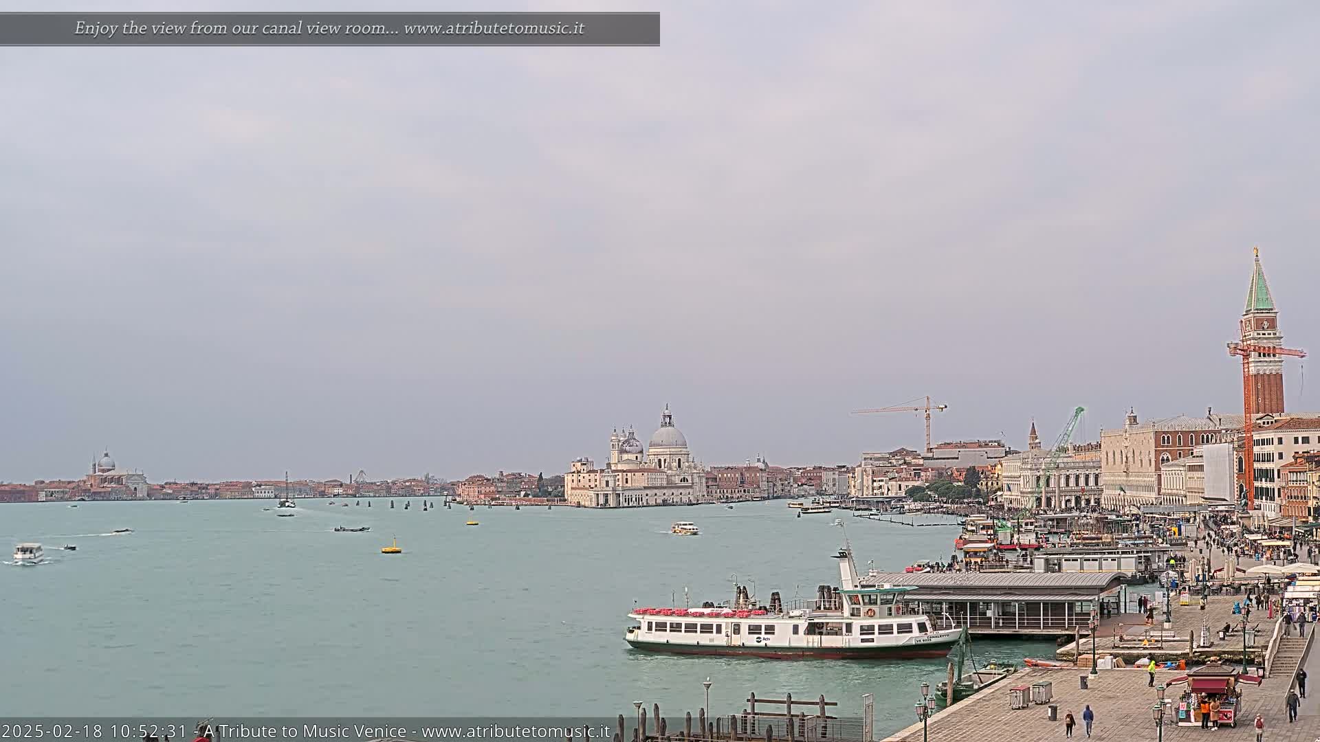 A wide shot of a Venetian canal on an overcast day shows a ferry docked near a waterfront walkway with several buildings in the background, including a large domed church and a tall bell tower.
