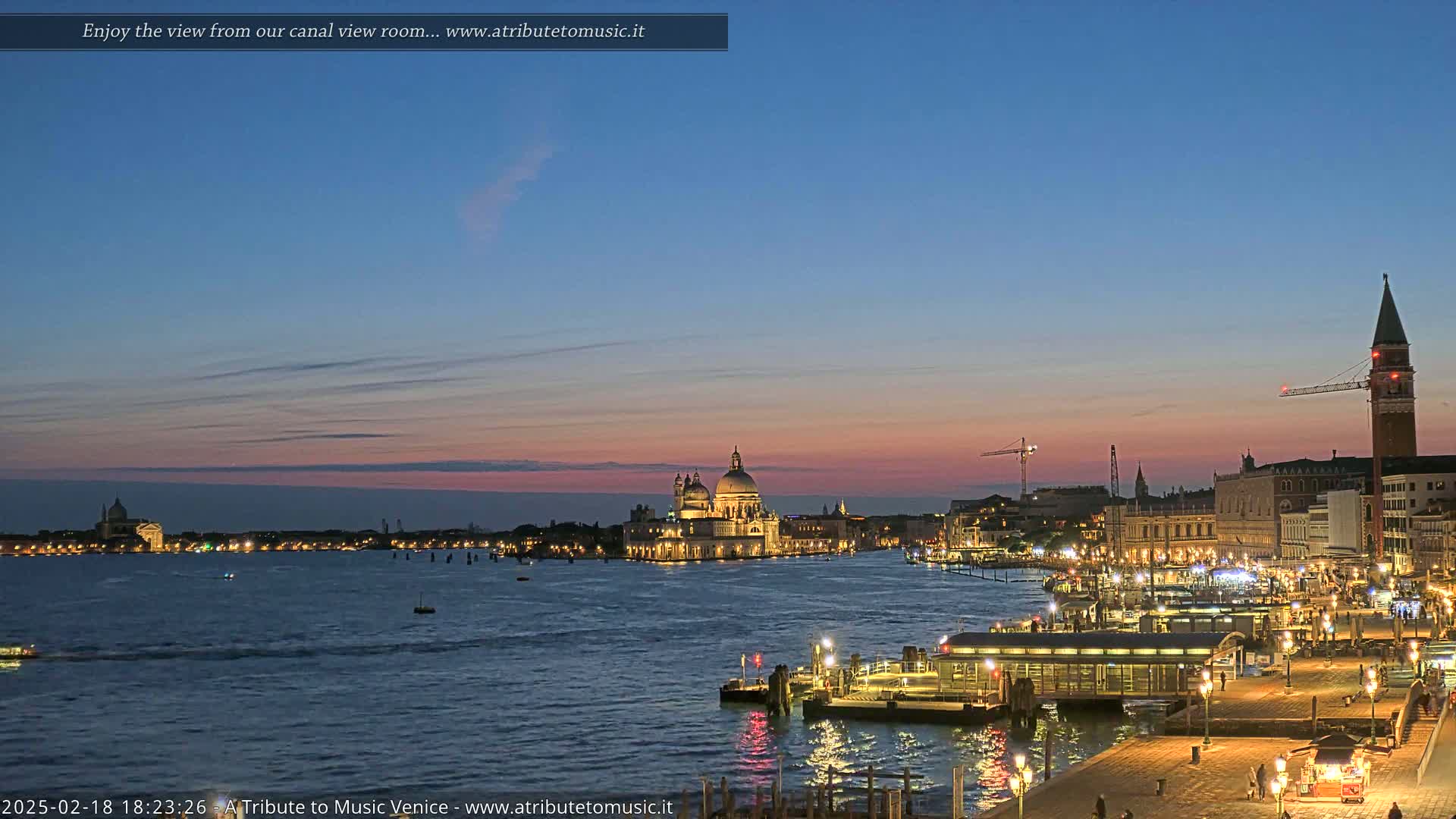 A twilight view of Venice, Italy, shows a calm canal with illuminated buildings and boats, under a dusky pink and blue sky.