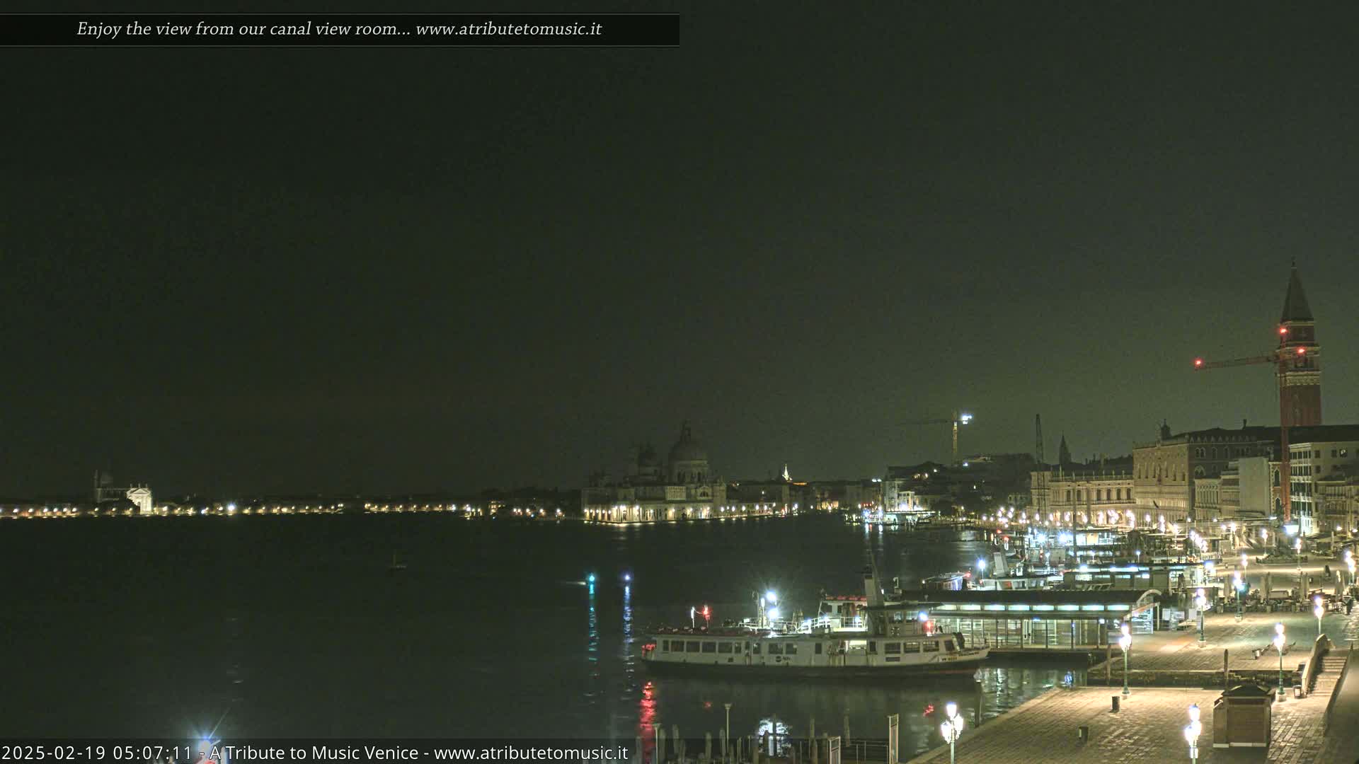 A nighttime view of a city waterfront with boats, buildings, and lights reflecting on the dark water under a clear, dark sky.