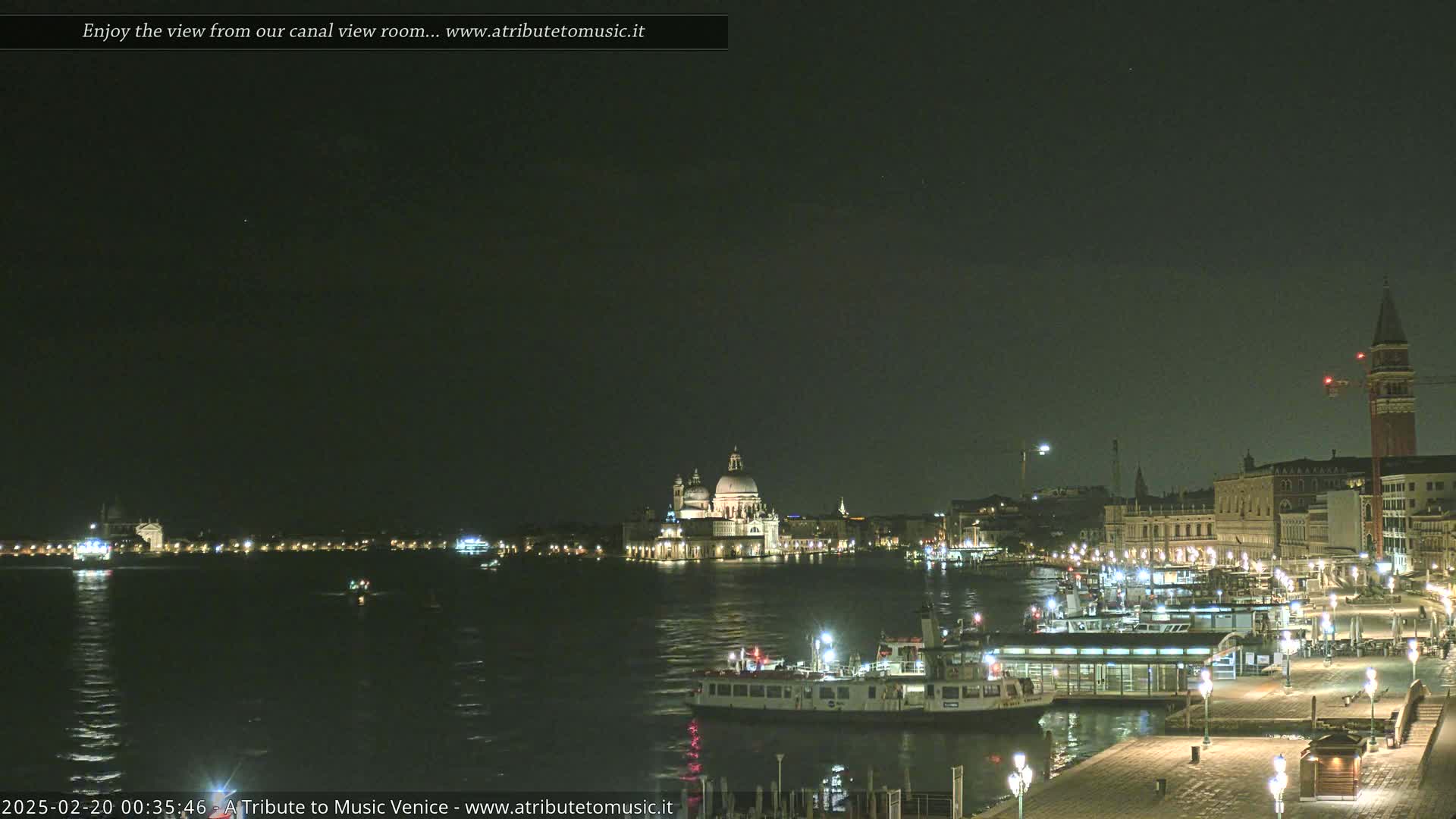 A nighttime view across a calm body of water shows illuminated buildings and boats in Venice under a clear, dark sky.