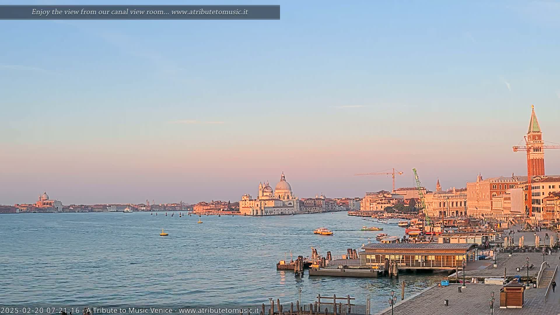 A wide, calm canal reflects the pale pink and blue hues of a sunrise sky, showcasing a cityscape of Venetian architecture, including a large domed church and a tall bell tower.