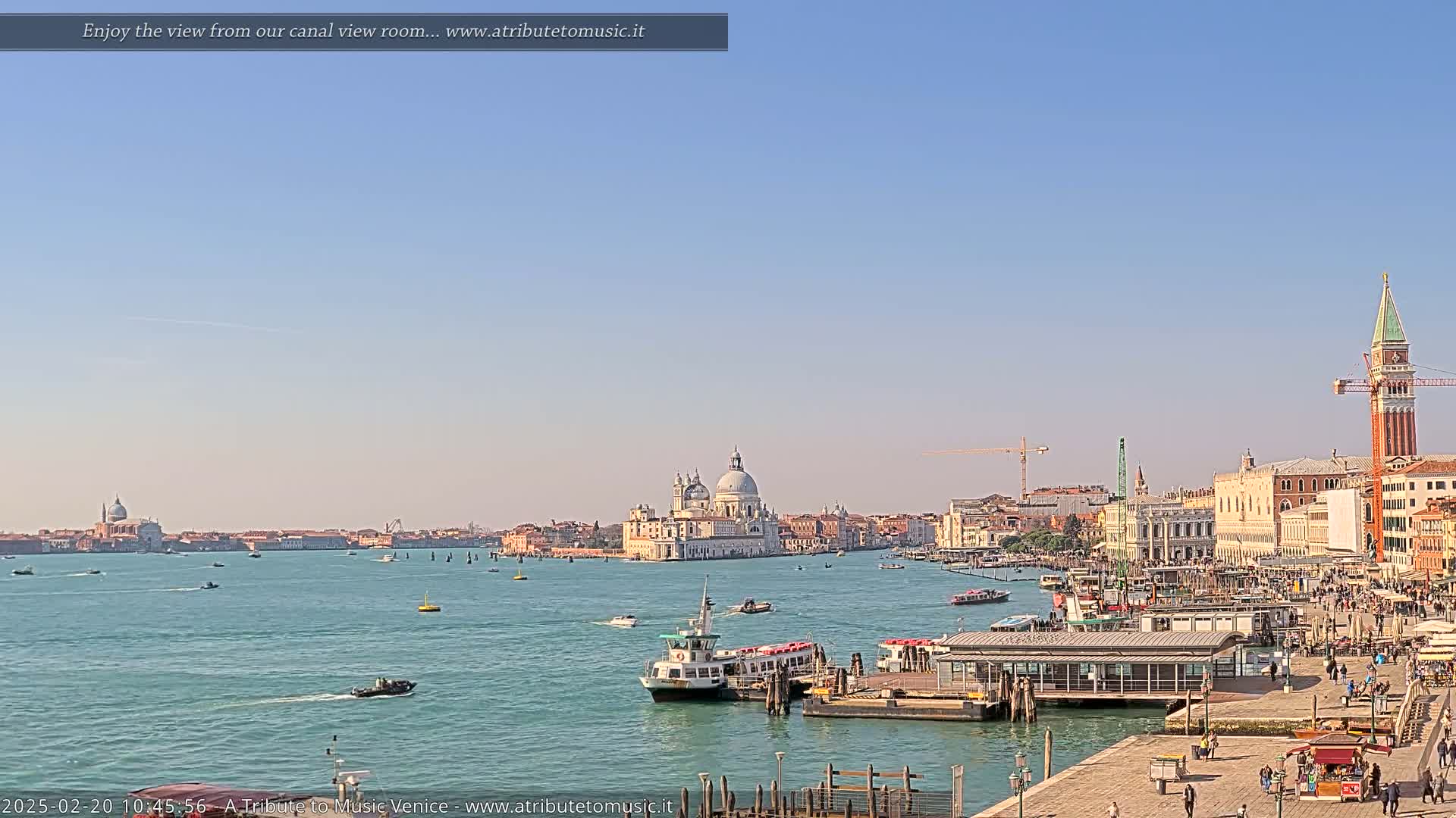 A sunny day in Venice shows a wide canal with boats and buildings along the shoreline, including a large domed church.