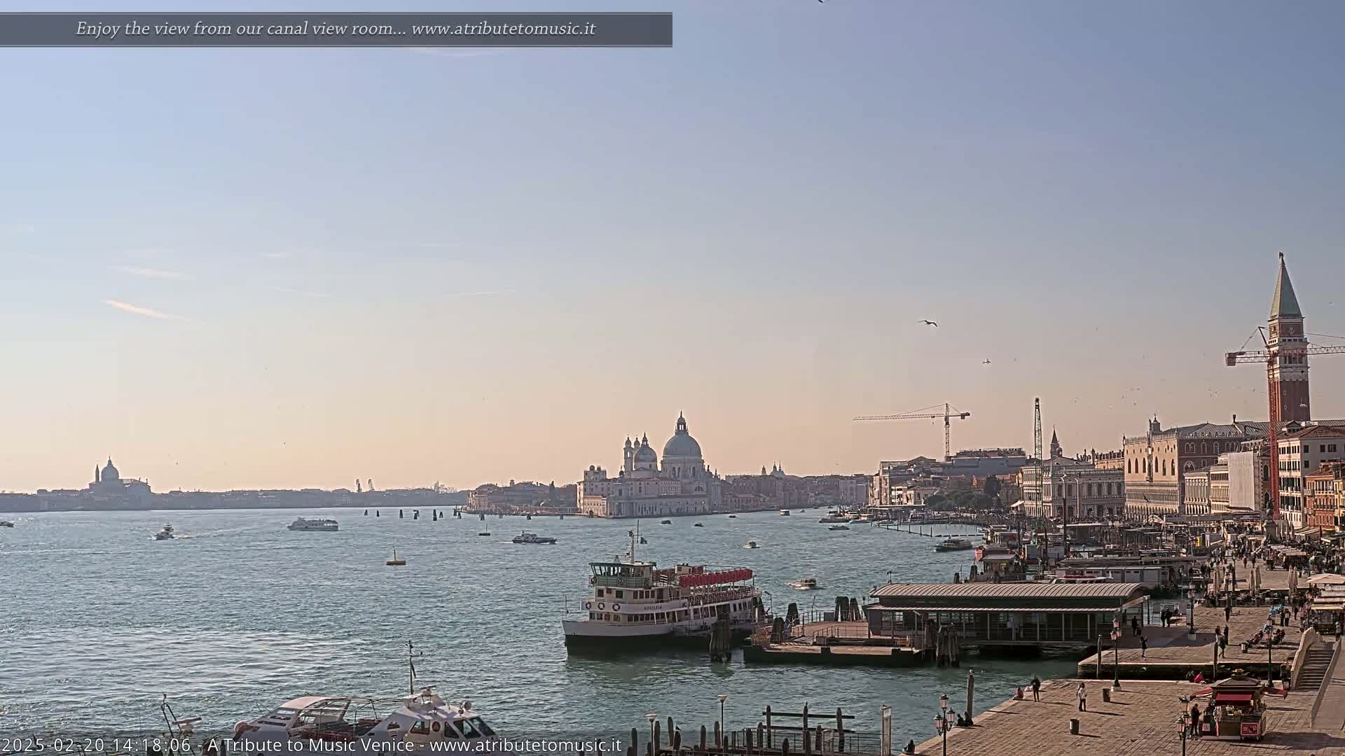 A calm waterway shows several boats, a large passenger ferry, and a cityscape under a clear, light-blue sky.