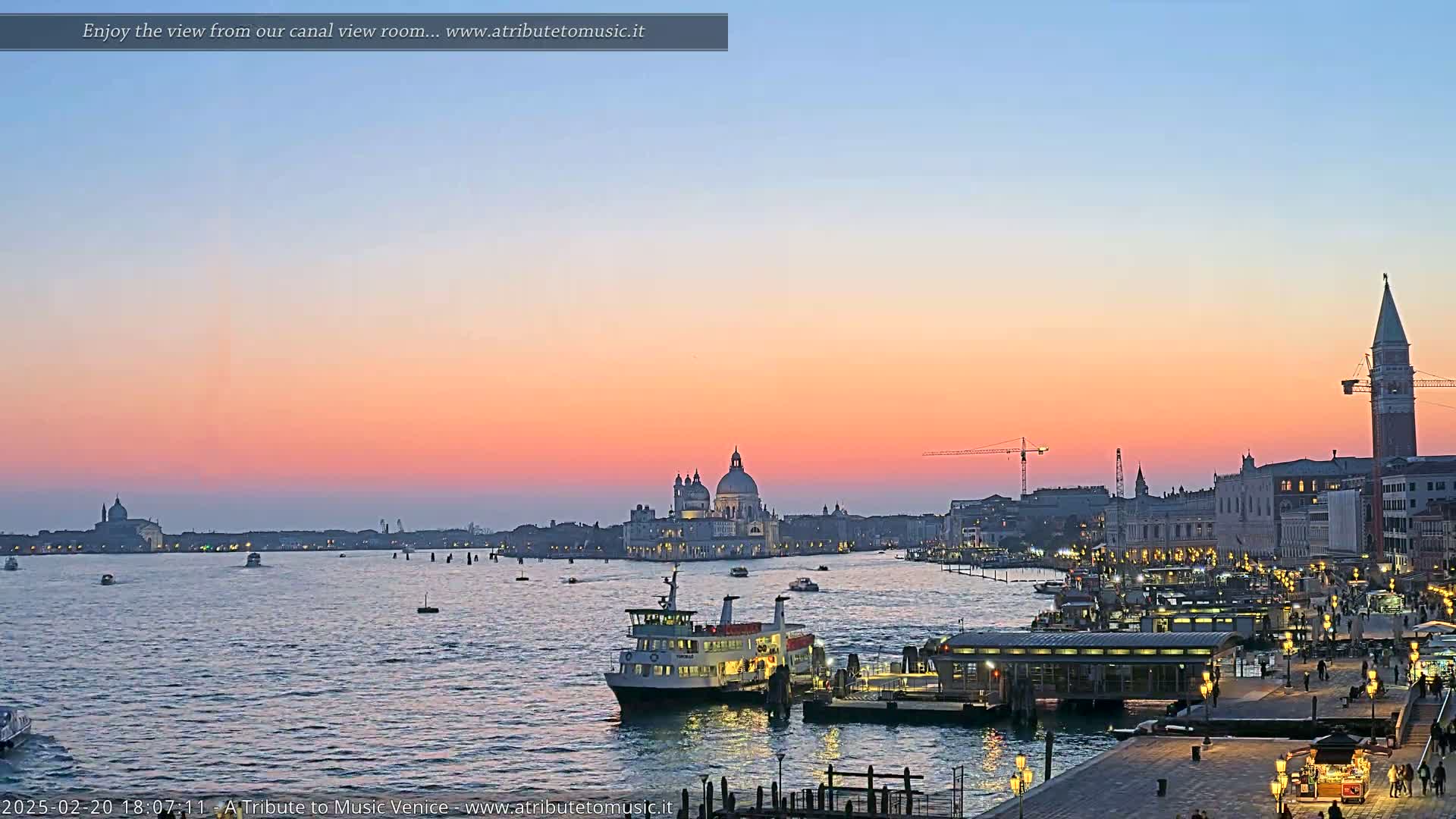 A Venetian waterway at sunset, with a ferry docked near a waterfront building, shows a cityscape with a large church dome in the distance under a pale pink and orange sky.