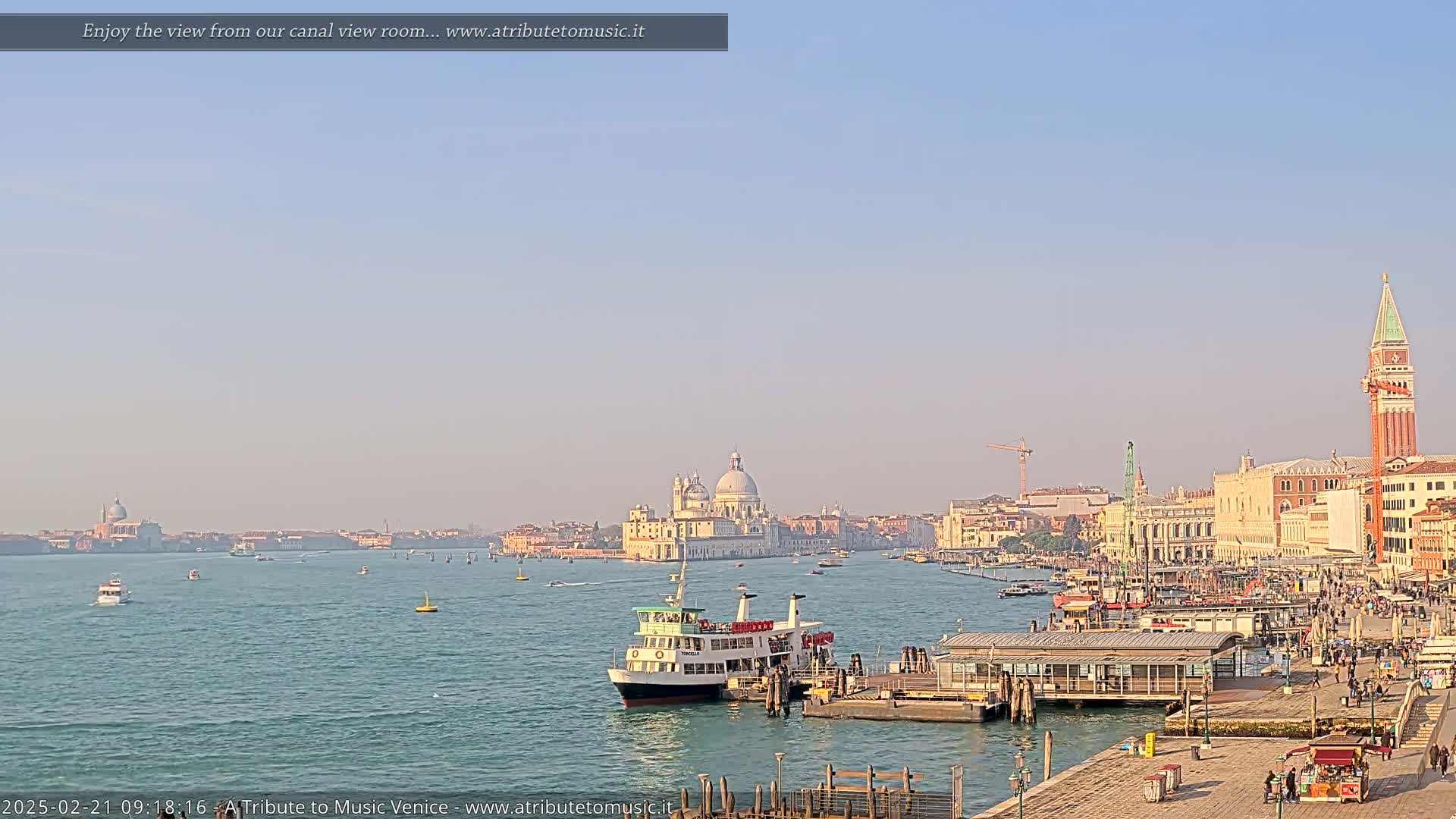 A ferry approaches a dock in a Venetian canal under a clear, light blue sky, with the Basilica di Santa Maria della Salute and St. Mark's Campanile visible in the distance.
