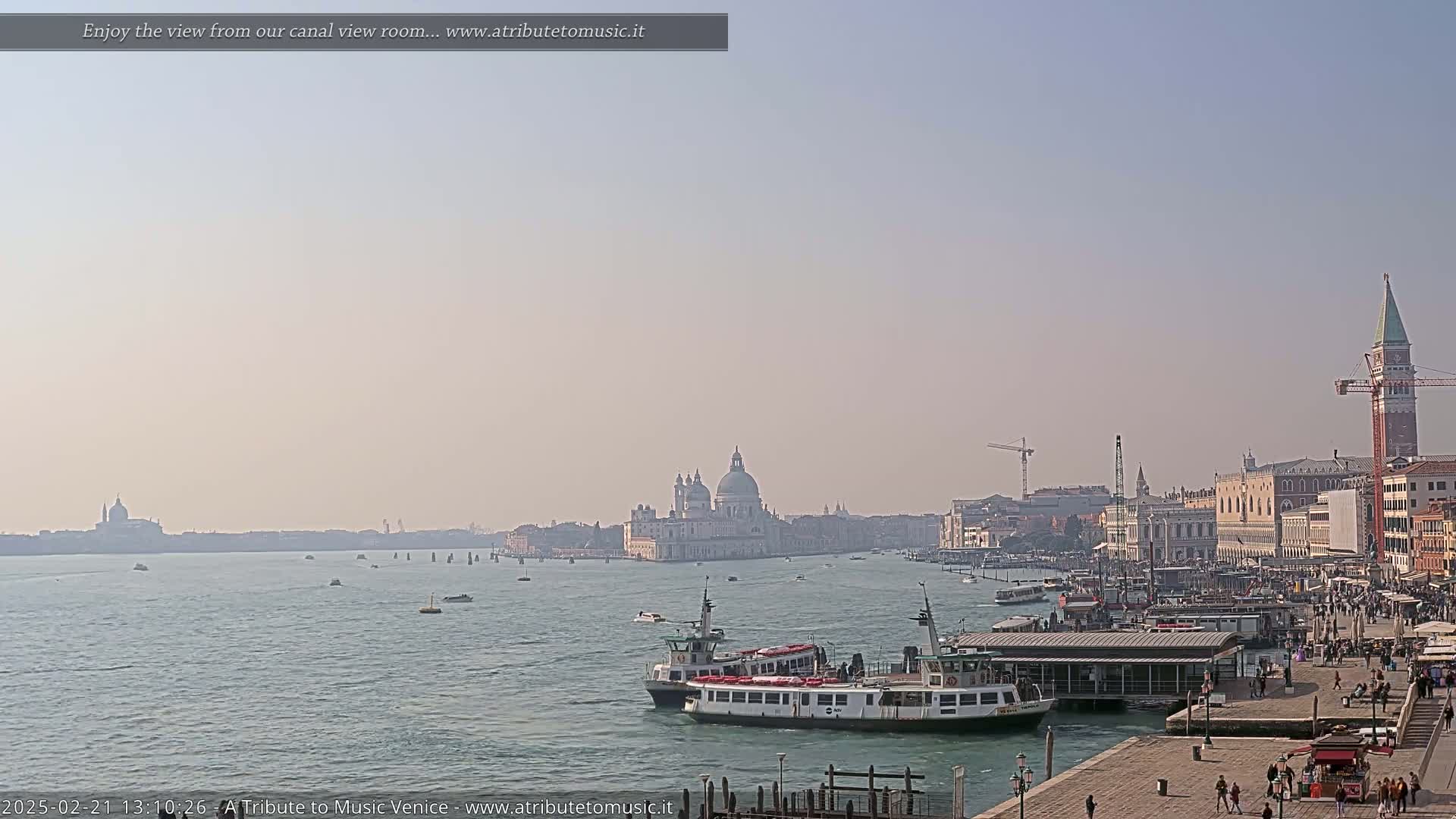 A hazy sunny day shows a wide Venetian canal with several boats, buildings lining the waterway, and a large domed church in the distance.