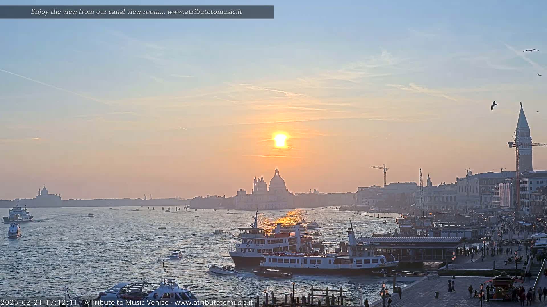 A hazy sunset over a Venetian canal, with several boats and the city skyline in the background.