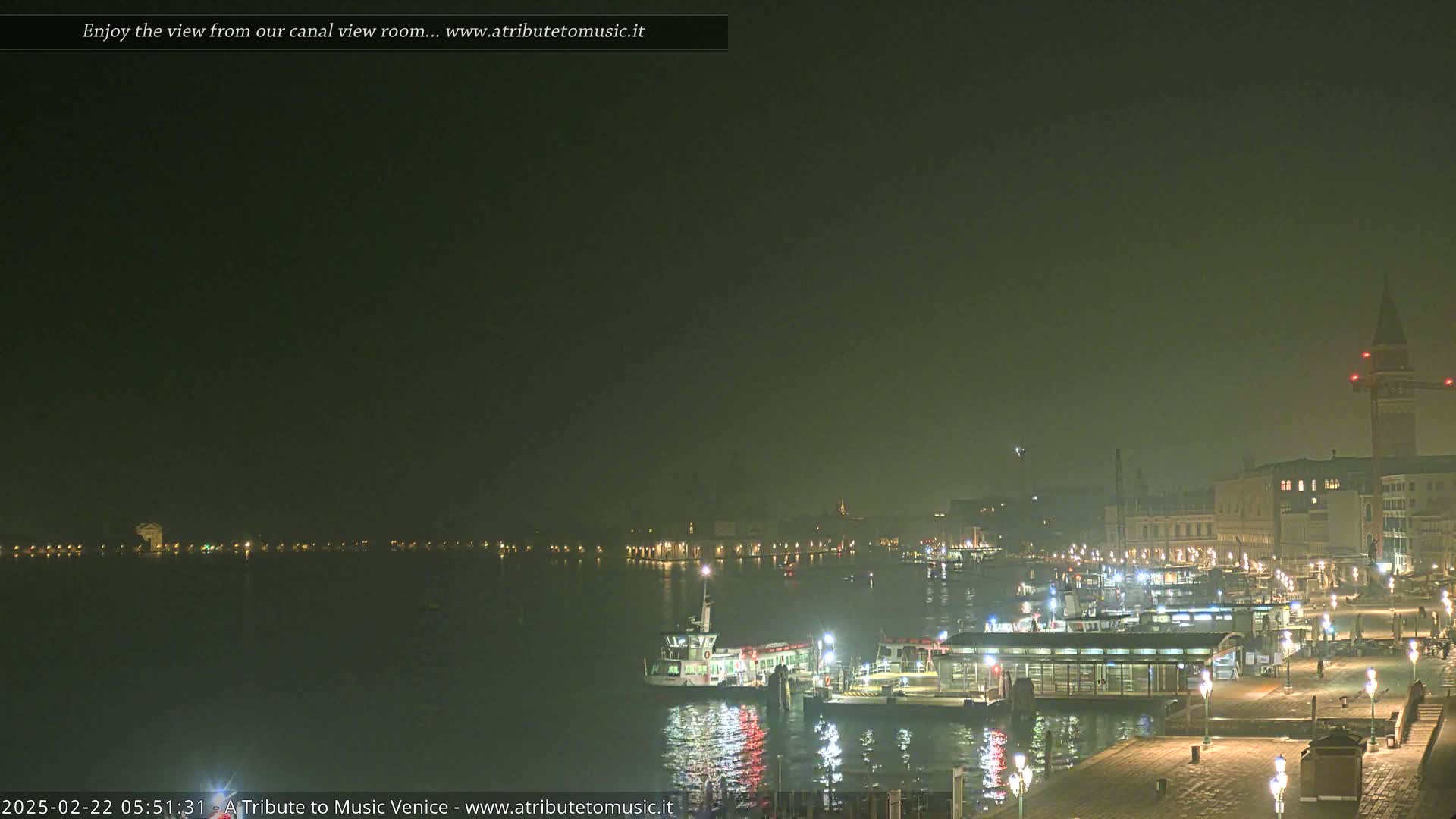 A nighttime view of a canal in Venice, shrouded in fog, shows a ferry and a waterfront with buildings and lights.