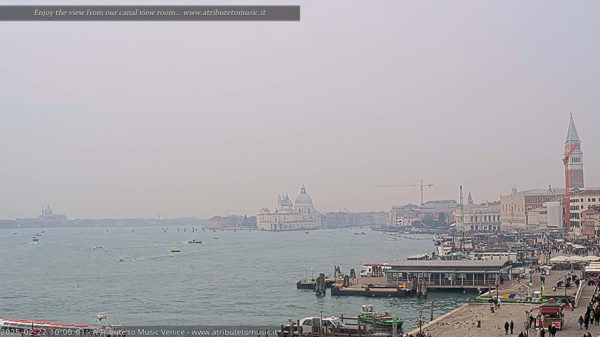 A hazy view across a canal shows a city with several buildings, boats, and people, under a light gray sky.