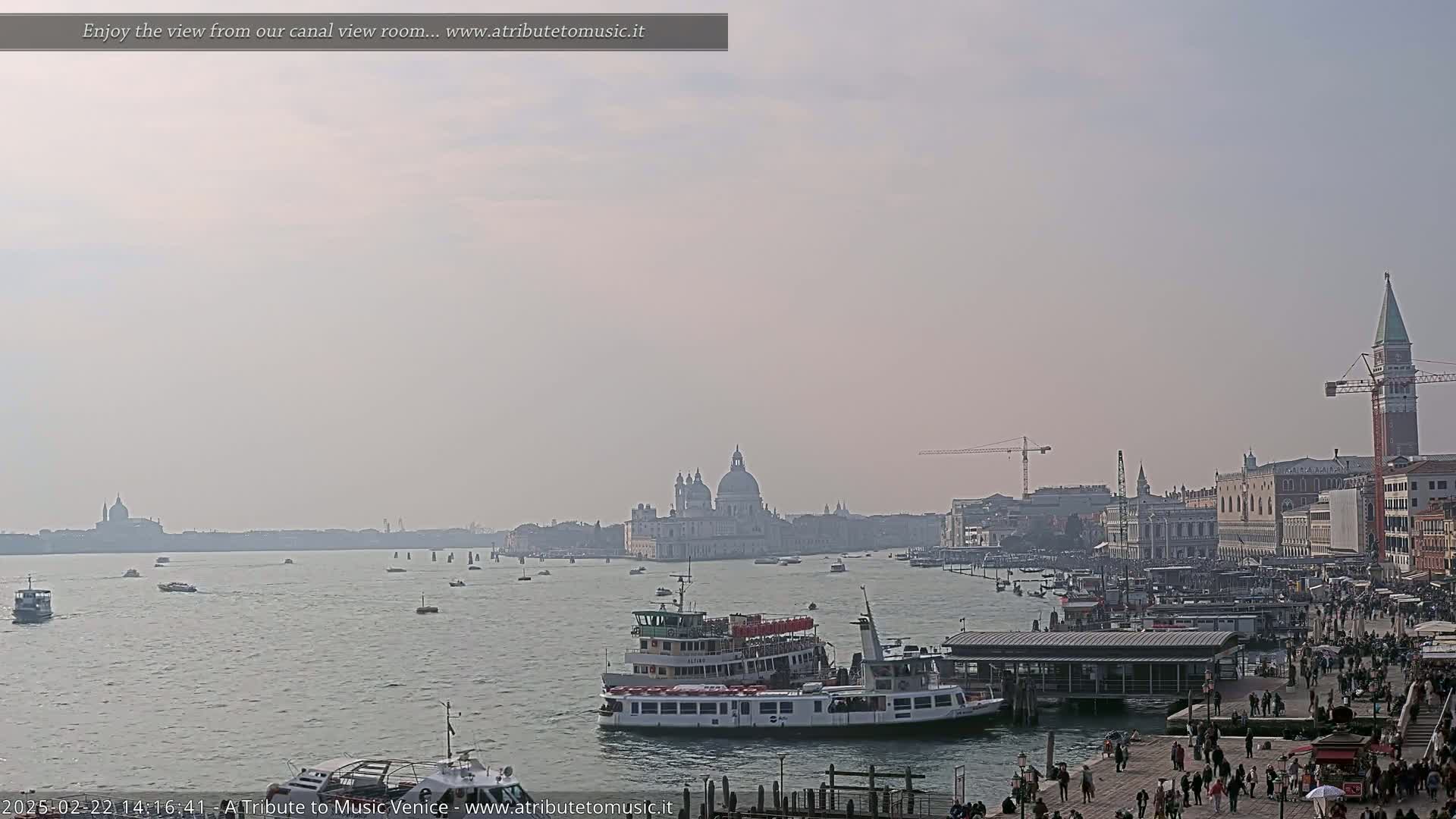 A hazy, sunny day in Venice shows a wide canal with several boats, a water taxi at a dock, and a distant view of the city including the Basilica di Santa Maria della Salute and St. Mark's Campanile.