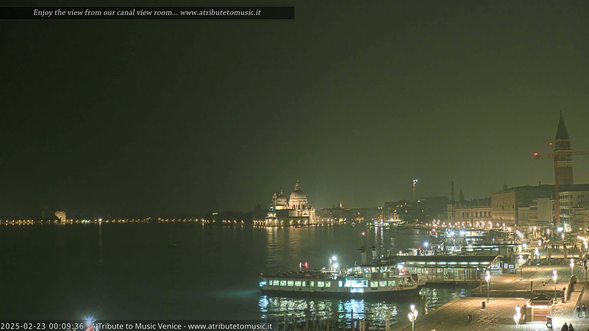 A nighttime view of a canal in Venice, showing a ferry docked near illuminated buildings and the Basilica di Santa Maria della Salute across the water under a dark sky.