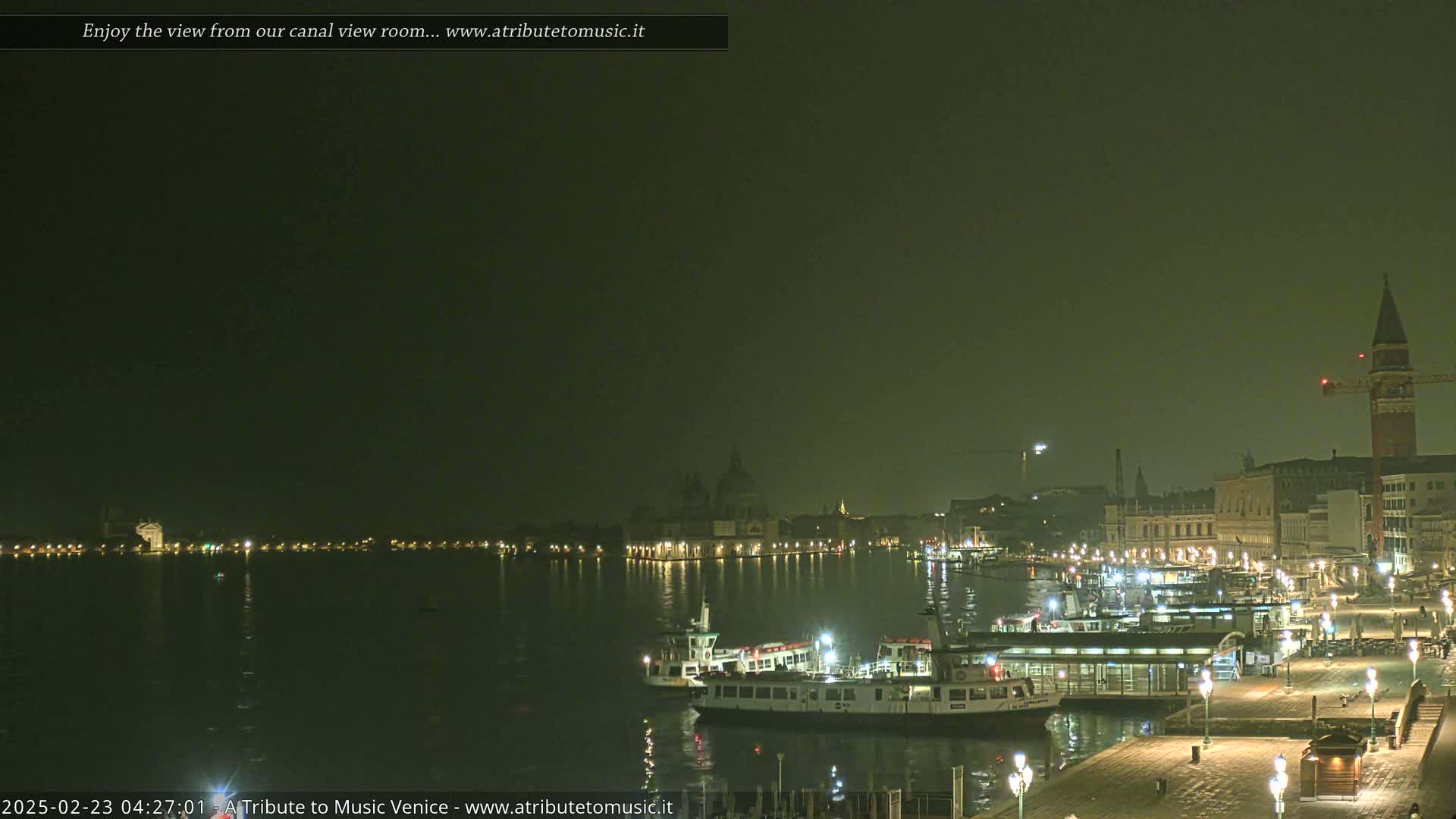 A nighttime view of a canal in Venice, Italy, shows boats docked along a waterfront with buildings and lights visible across the water under a hazy sky.