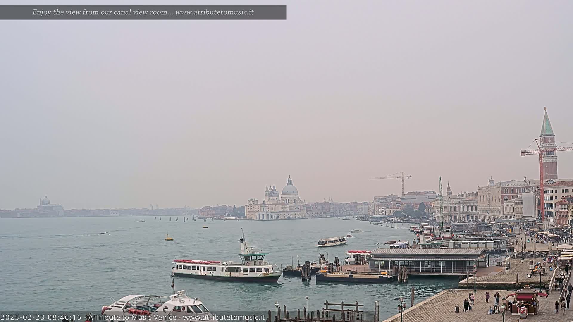 A hazy view across a canal shows several boats near a dock, with a large building and cityscape in the distance.