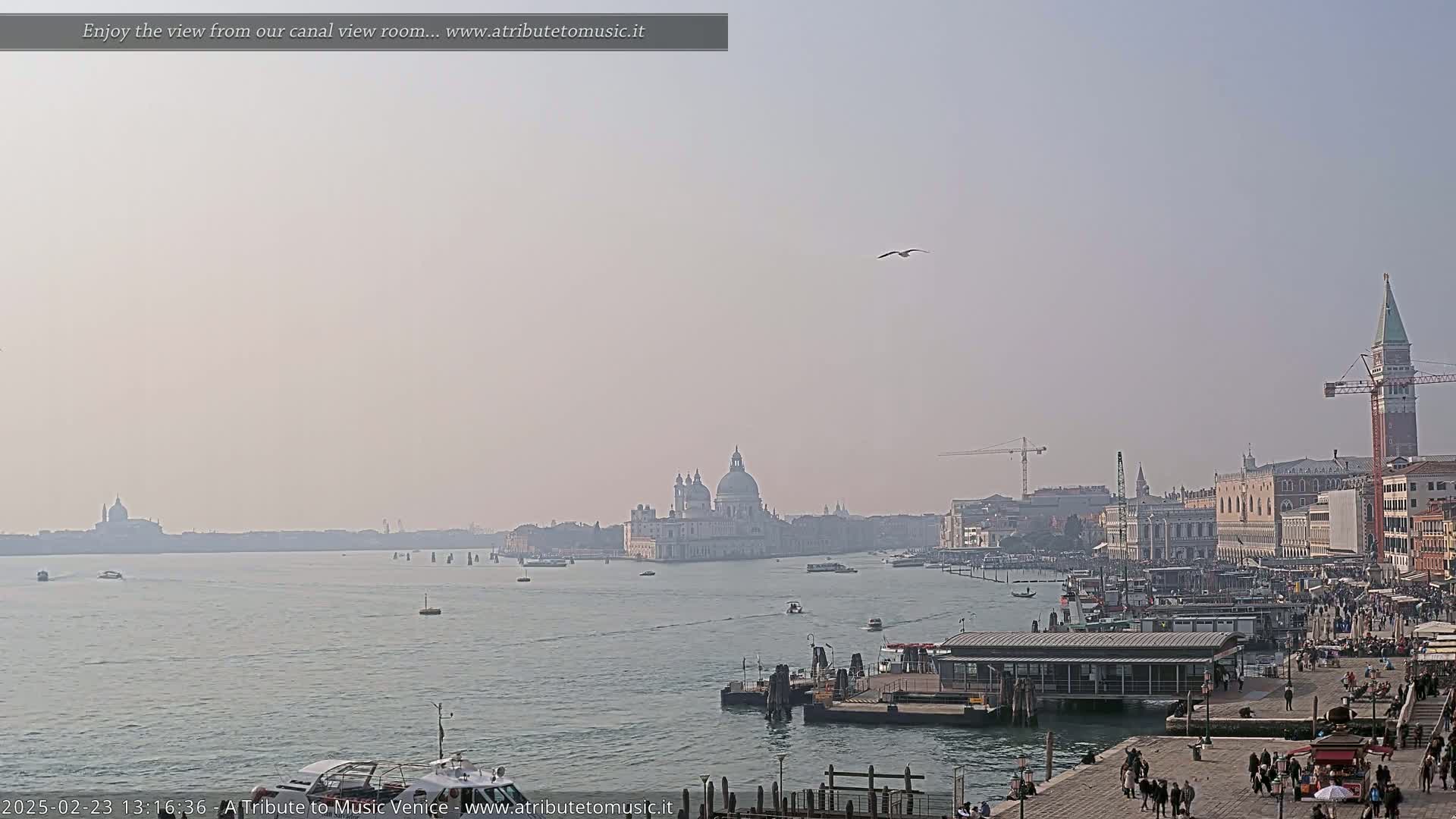 A hazy, sunny day reveals a wide canal view of Venice, Italy, showcasing the Basilica di Santa Maria della Salute and other buildings along the waterfront, with boats and people visible.