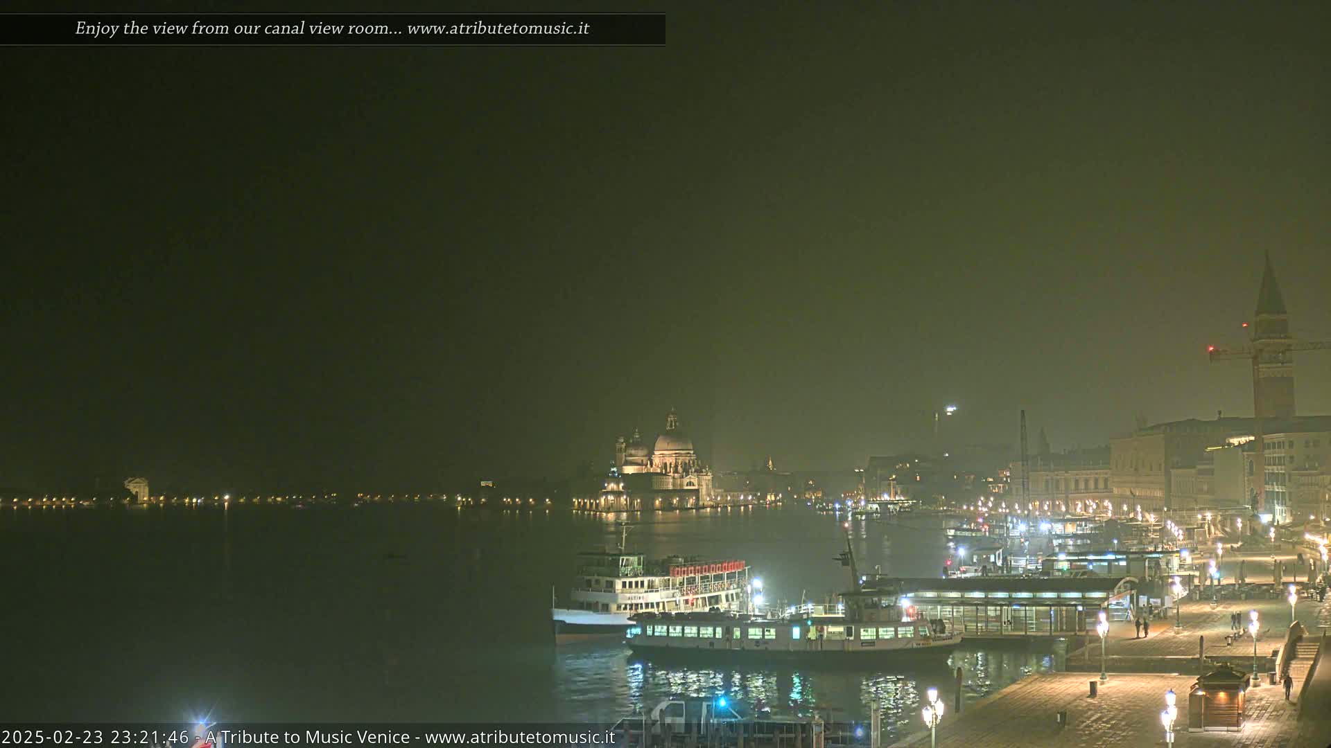 A nighttime view of a canal in Venice, shrouded in fog, shows docked boats and illuminated buildings across the water.