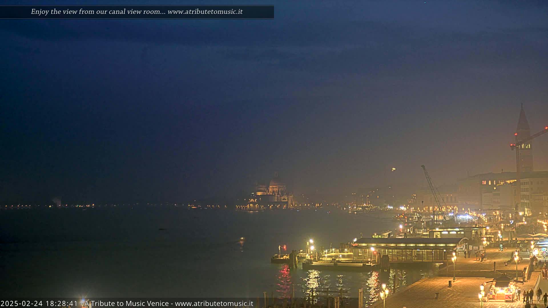 A hazy, twilight view of a city waterfront at night, showing a calm body of water with lights along a dock and buildings in the distance, including a domed structure.