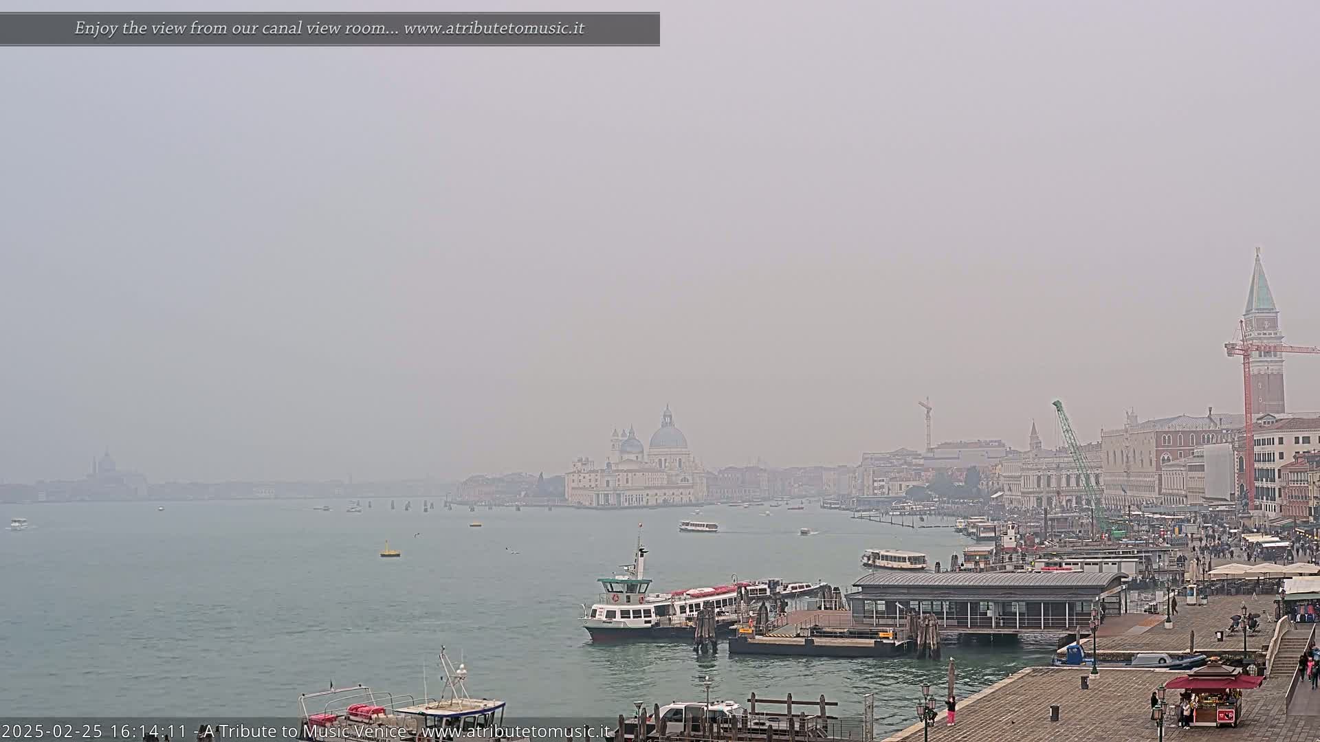 A hazy view of a Venetian canal shows several boats docked near buildings, with the Basilica di Santa Maria della Salute visible in the distance.