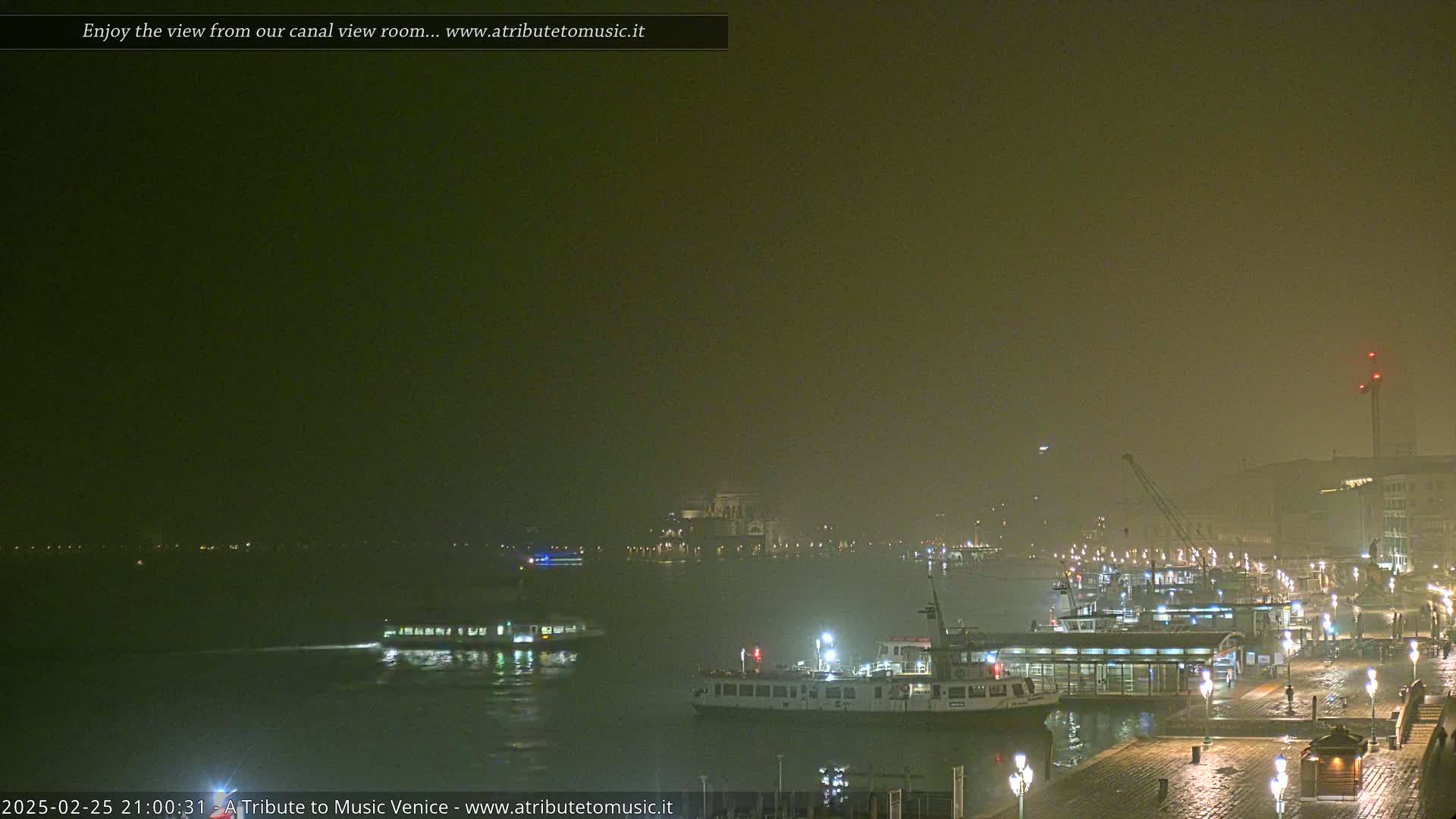 A nighttime view of a canal with several boats and a hazy atmosphere, showing lights along the waterfront.